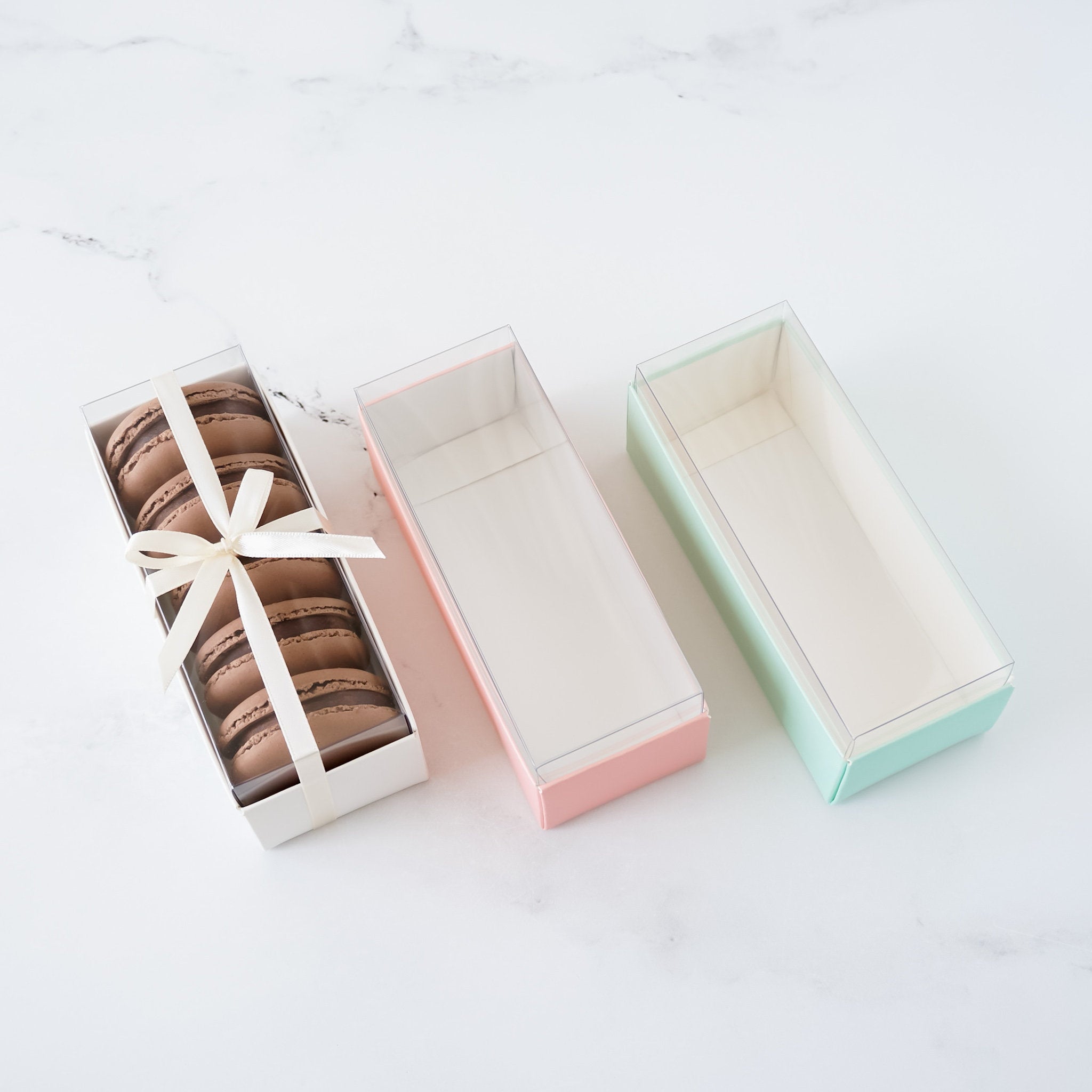 Three clear plastic boxes with lids, one is white, one is pink, and one is light green. The white box contains six chocolate macarons tied with a white ribbon.