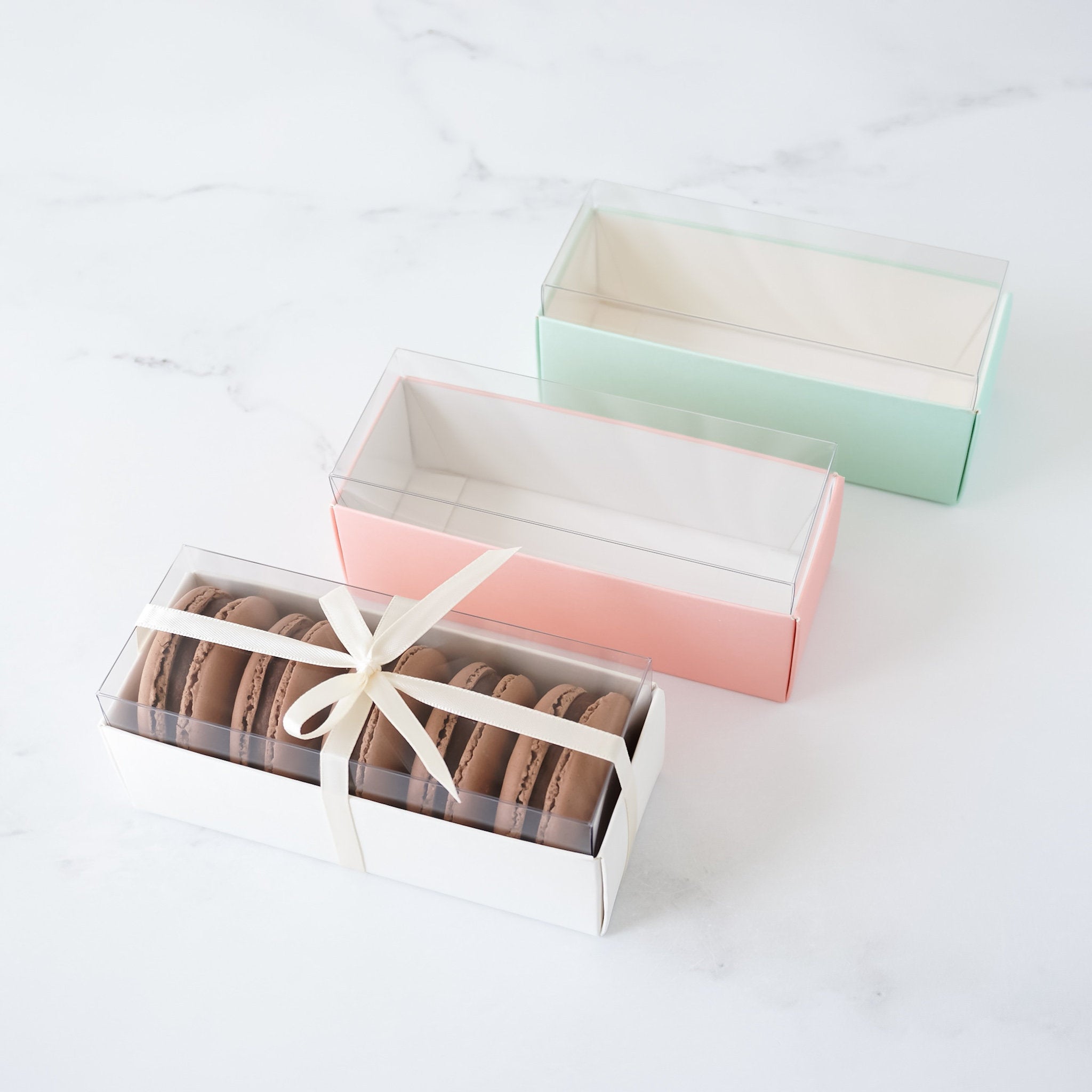 Three rectangular boxes with clear lids. The top box is light green, the middle box is light pink, and the bottom box is white. The bottom box contains six chocolate macarons tied with a white ribbon.