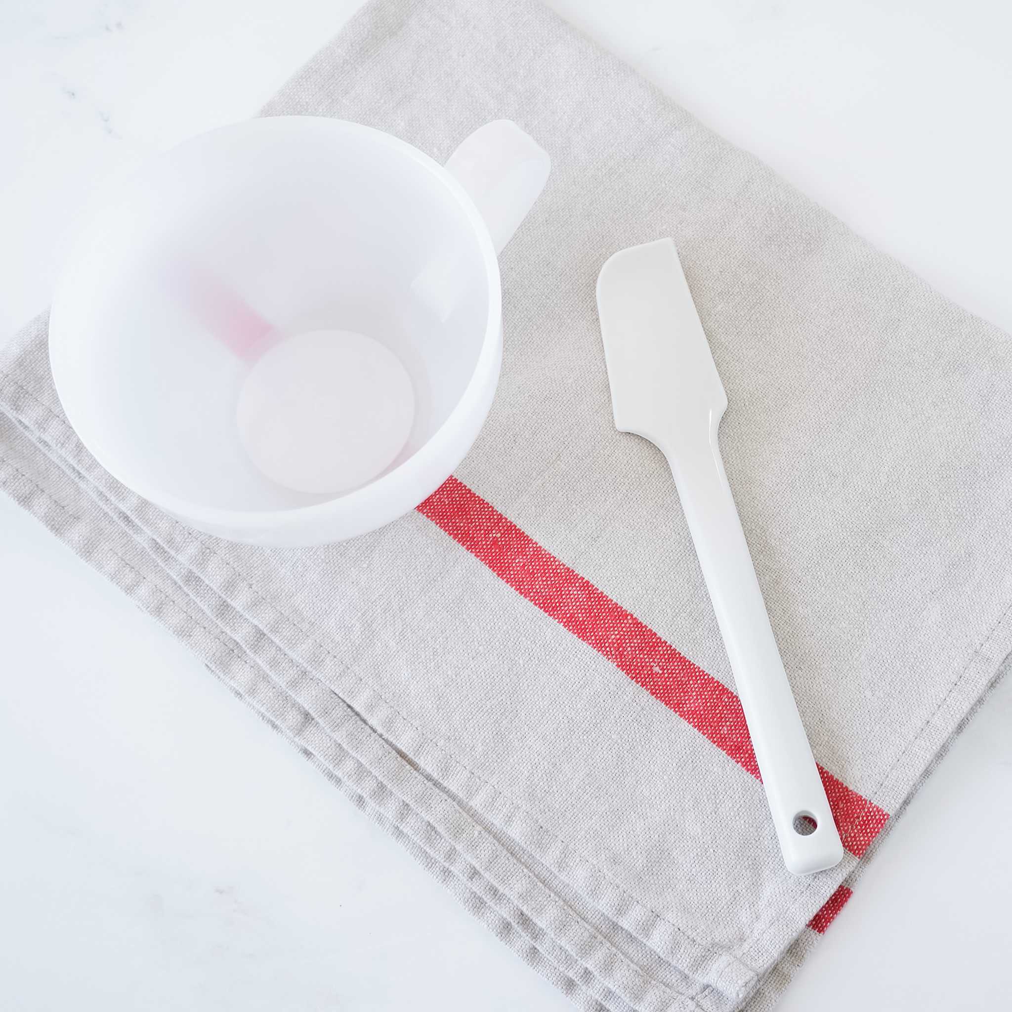 A white mixing bowl with a handle and a white silicone spatula on a folded linen kitchen towel with a red stripe.
