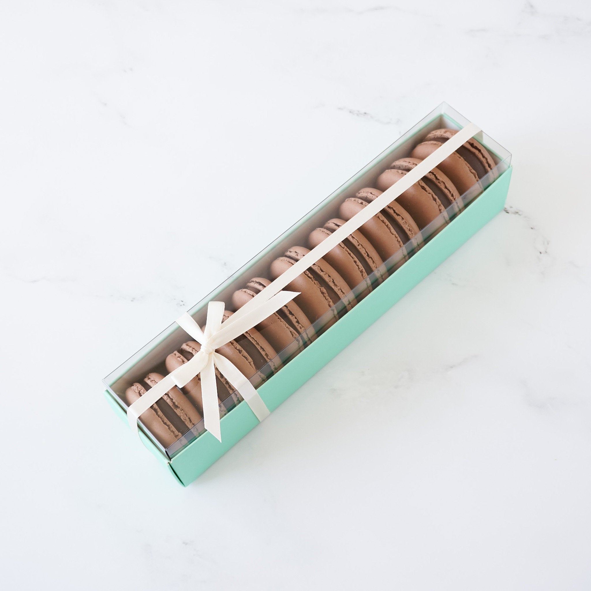 A box of chocolate macarons tied with a white ribbon. The box is light green and the macarons are arranged in a row inside the box.