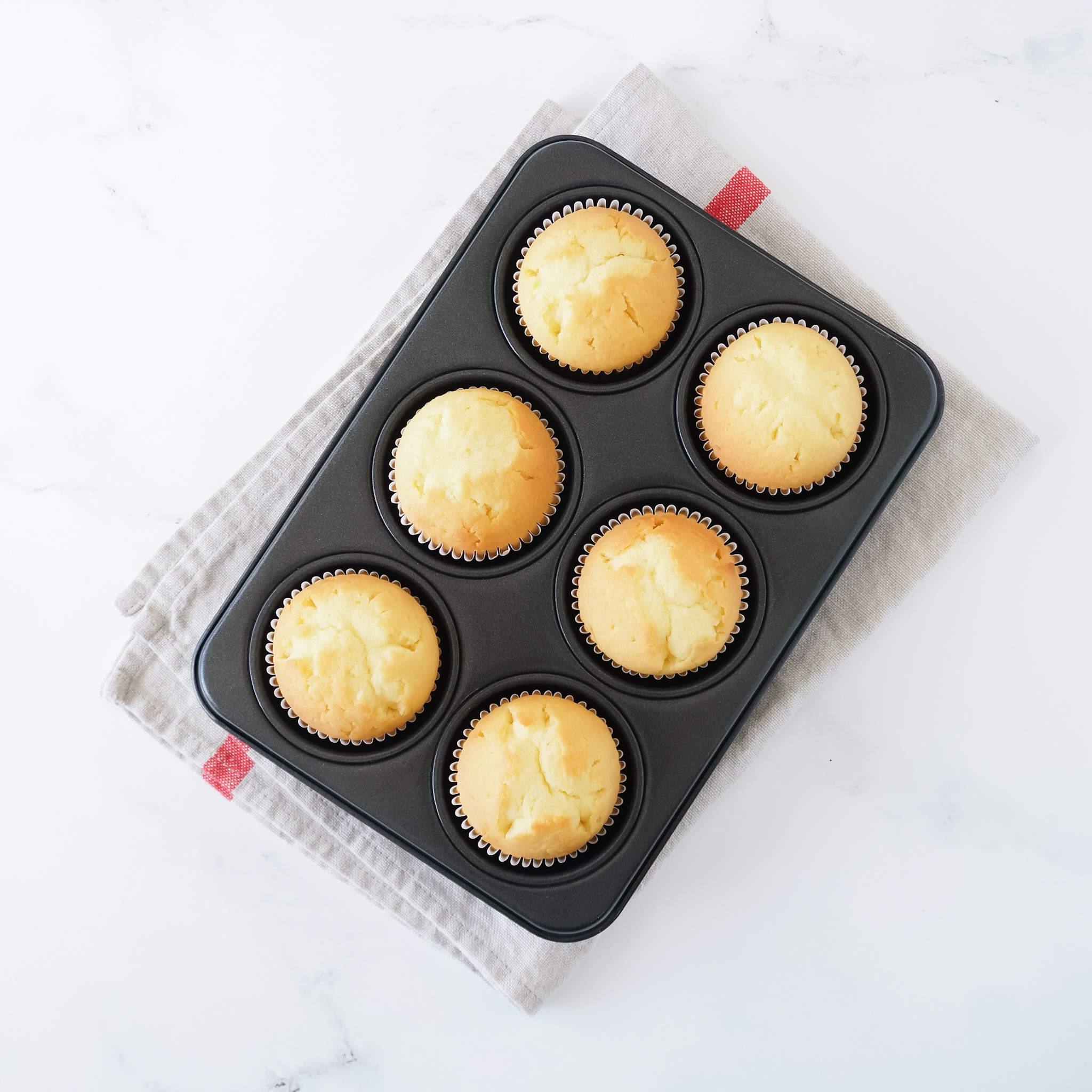 A 6-cup muffin and cupcake pan with golden-brown baked cupcakes inside, placed on a white surface with a light grey napkin at the bottom.