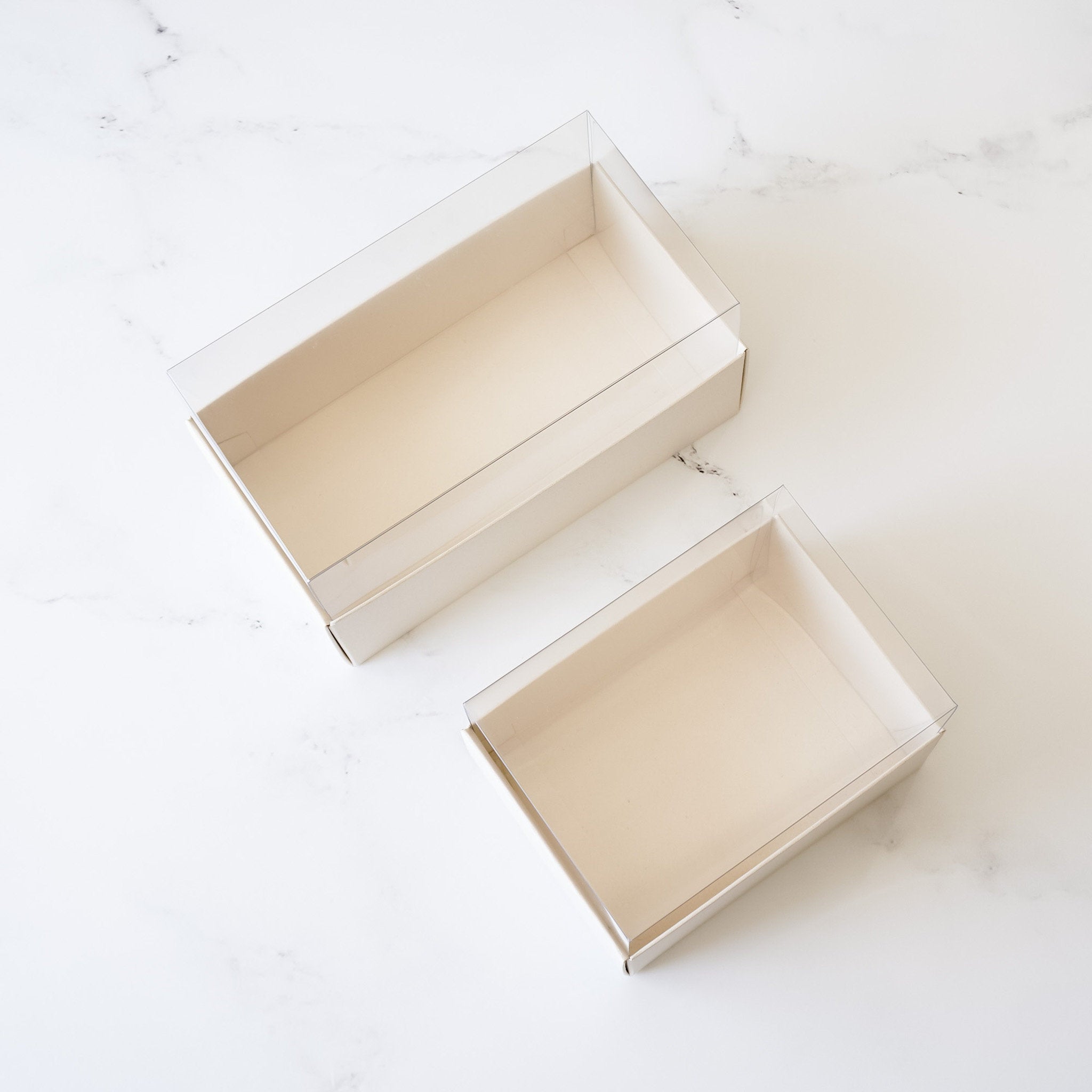 Two empty rectangular boxes with clear plastic lids. The boxes are made of light beige cardboard and are sitting on a white marble surface.