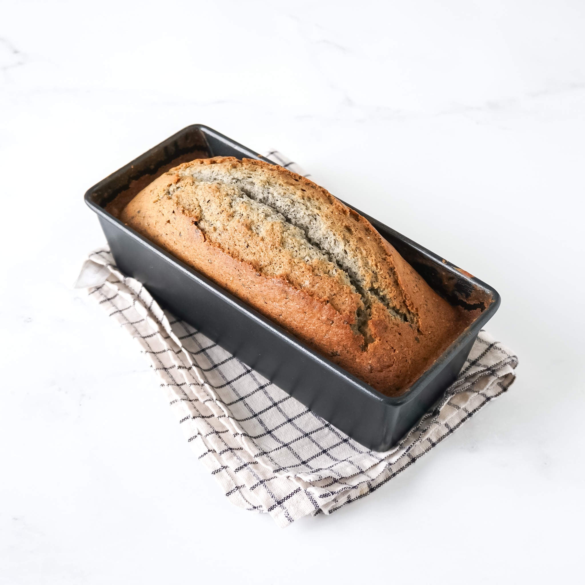 A golden-brown pound cake in a loaf pan, placed on a white surface with a checkered kitchen cloth.