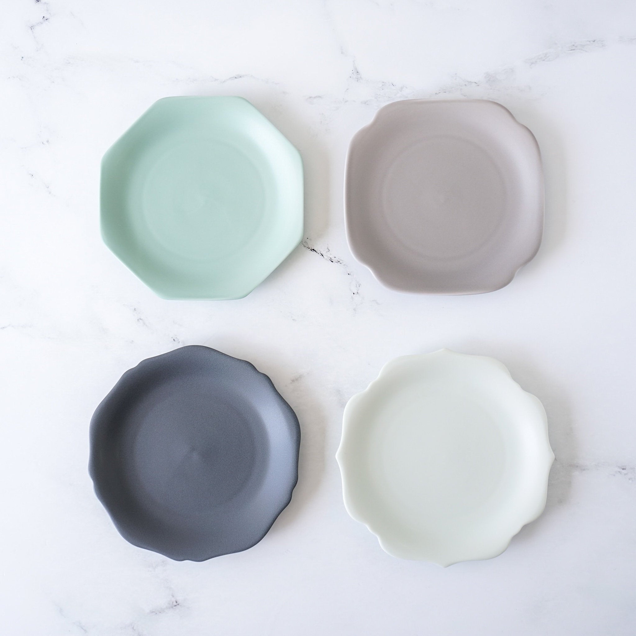 A set of four porcelain dessert plates in different colors including green, gray, blue, and white, displayed on a marble surface.