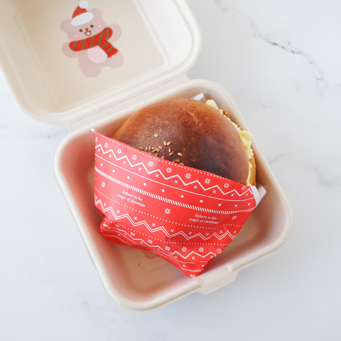 A festive red double open paper bag with a Christmas print, containing a sandwich, placed inside a take-out container.