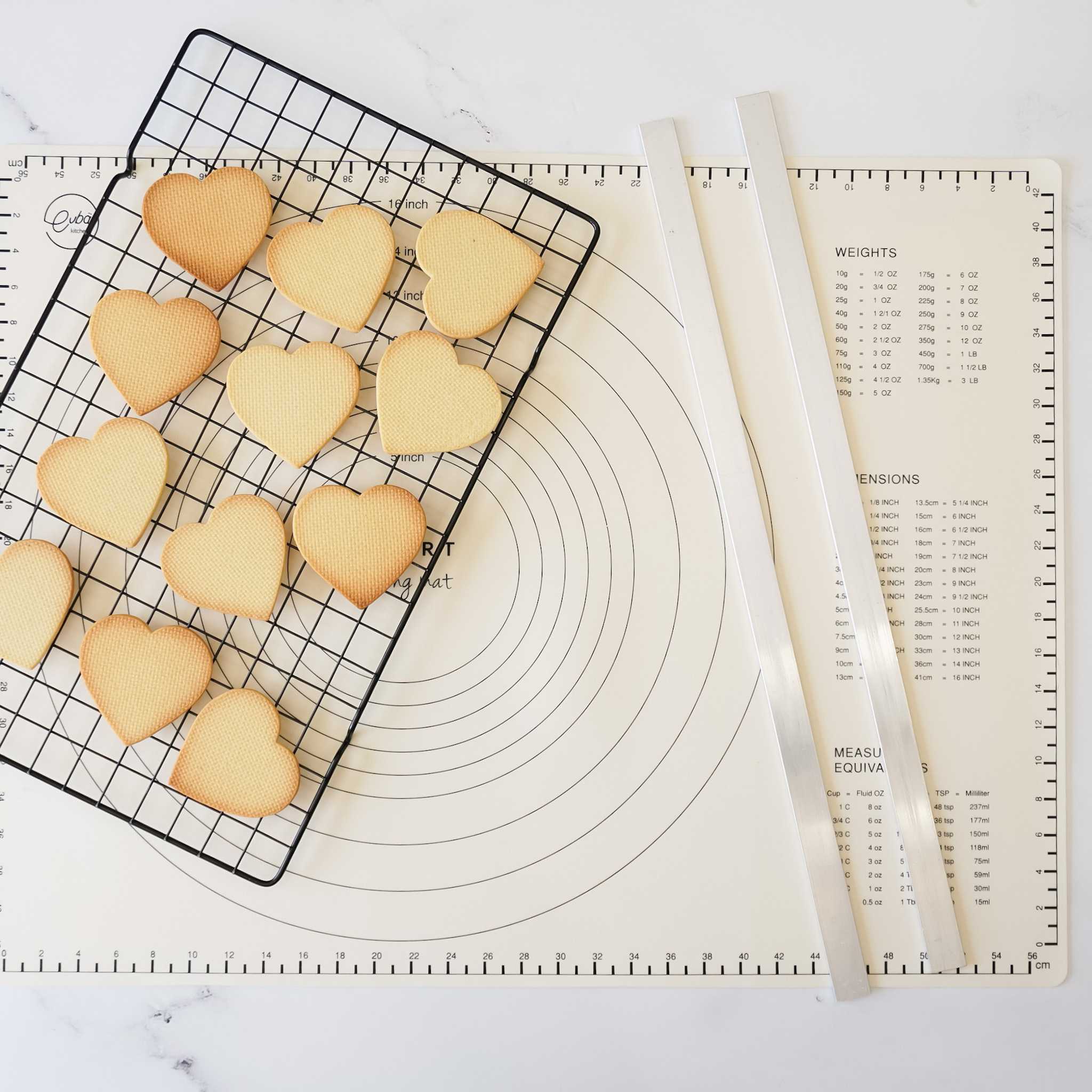 A set of two aluminum pastry rulers with measurement markings, alongside a cooling rack with heart-shaped cookies on it, placed on a white surface.