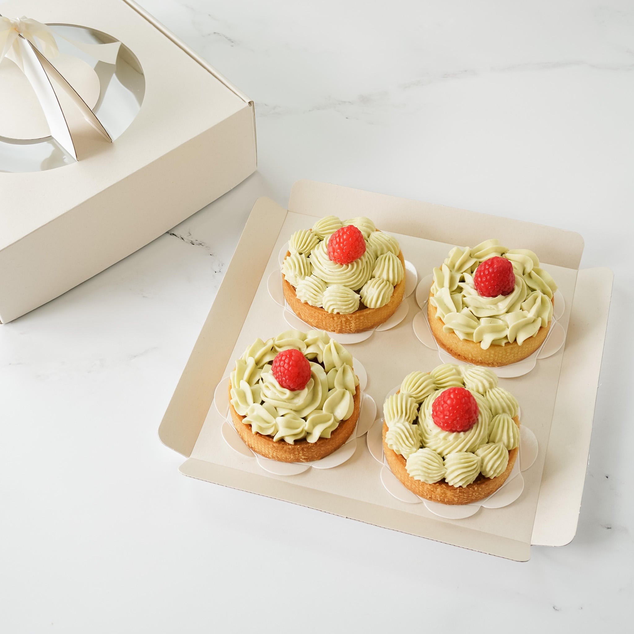 Four green and white frosted mini tarts with a single red raspberry on top, arranged in a light beige cardboard box.