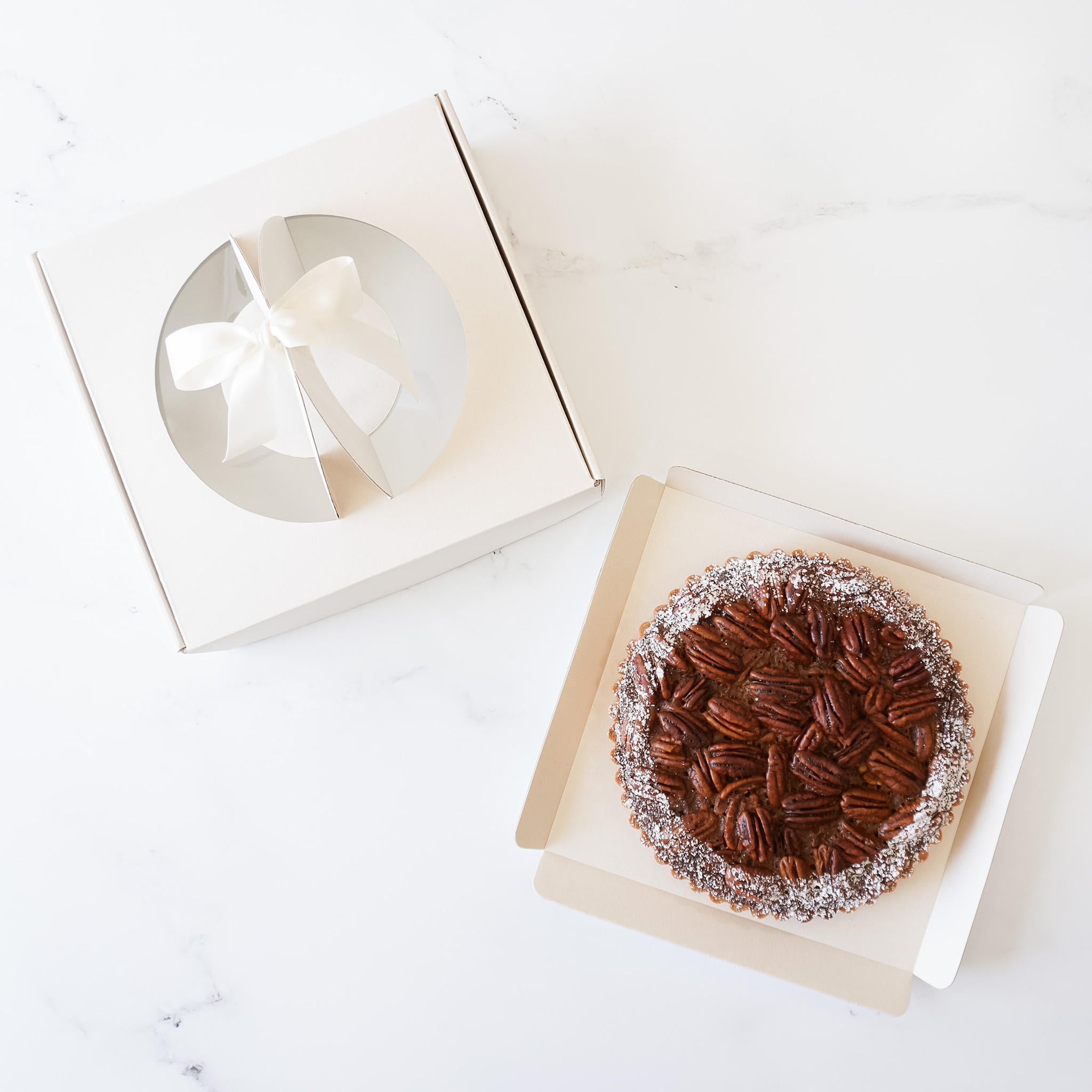 An unassembled square bakery box in a natural beige color with a clear window on top, containing a pie inside, placed on a white surface.