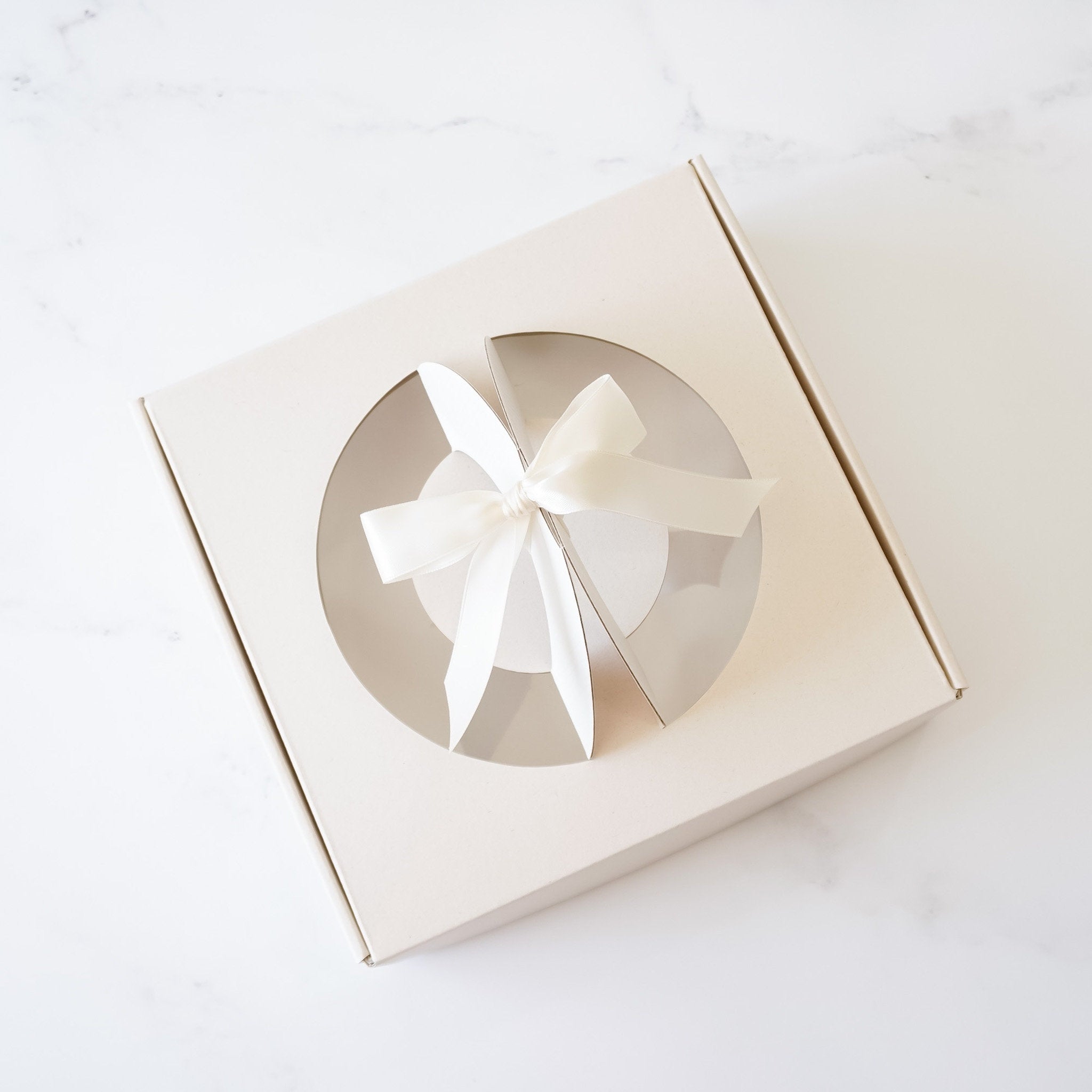 A light beige cardboard box with a circular window and a white satin ribbon tied in a bow. The box is designed to hold a cake or other baked good.