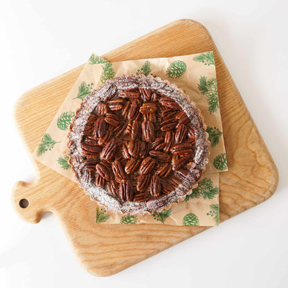 A round pecan pie or tart topped with whole pecans and dusted with powdered sugar, presented on a patterned parchment paper sheet over a wooden cutting board against a white background.