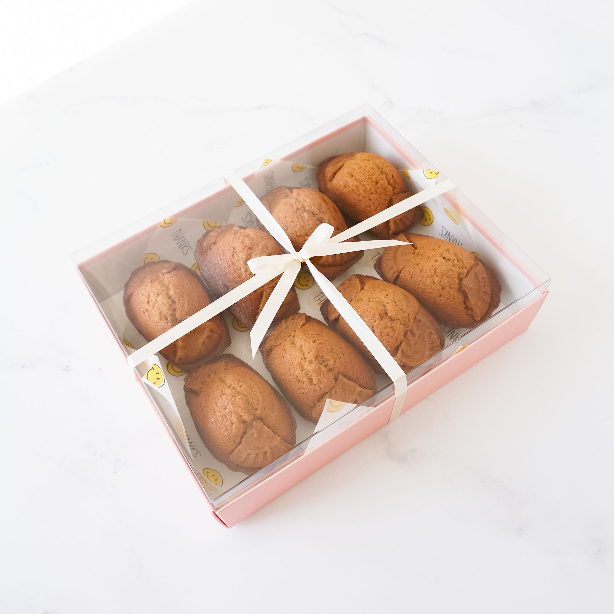 A pink box with a clear lid filled with eight small, brown, baked goods. The box is tied with a white ribbon and bow. The box has a white and yellow patterned paper lining with the words &