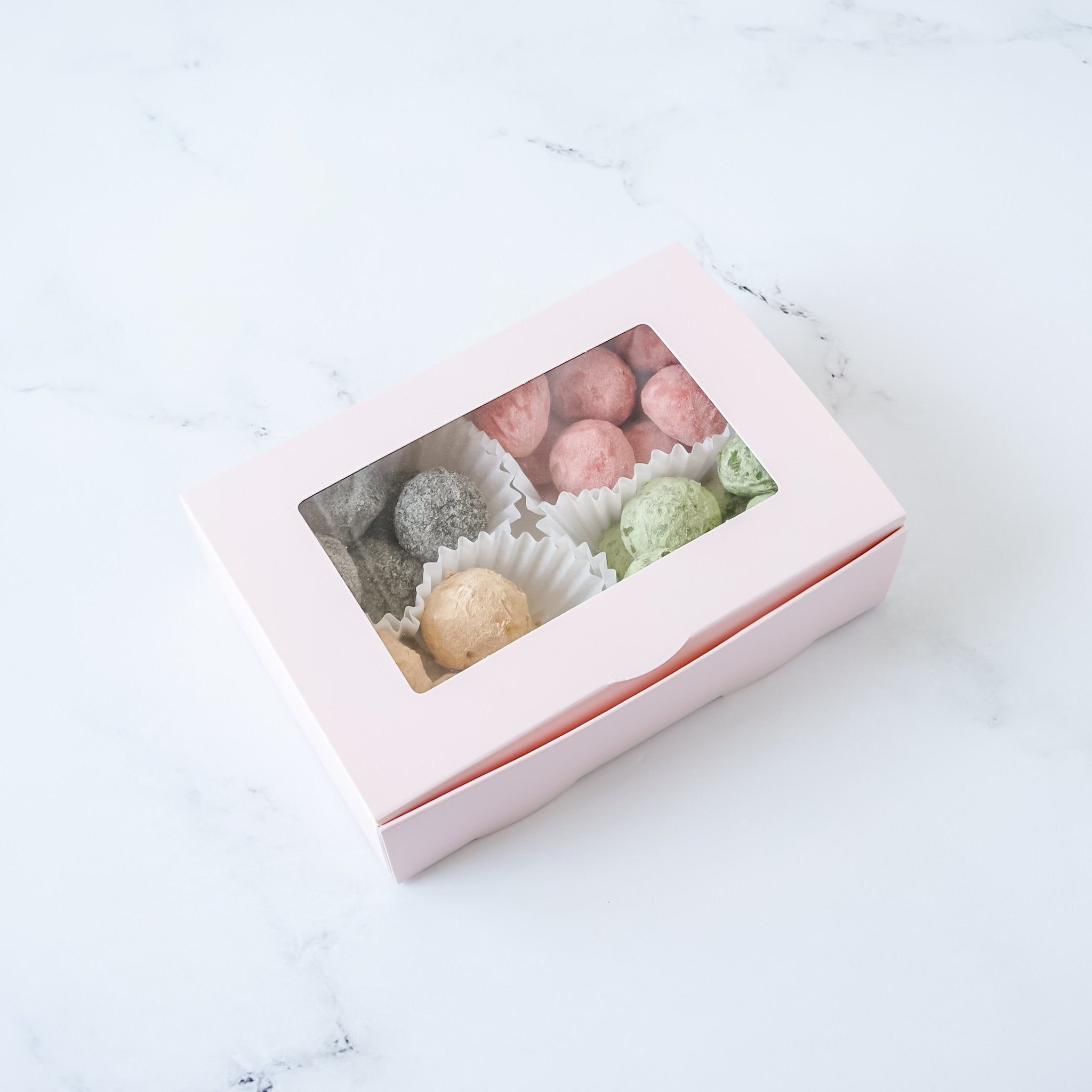 A blush pink cookie box with a clear window displaying various cookies inside.