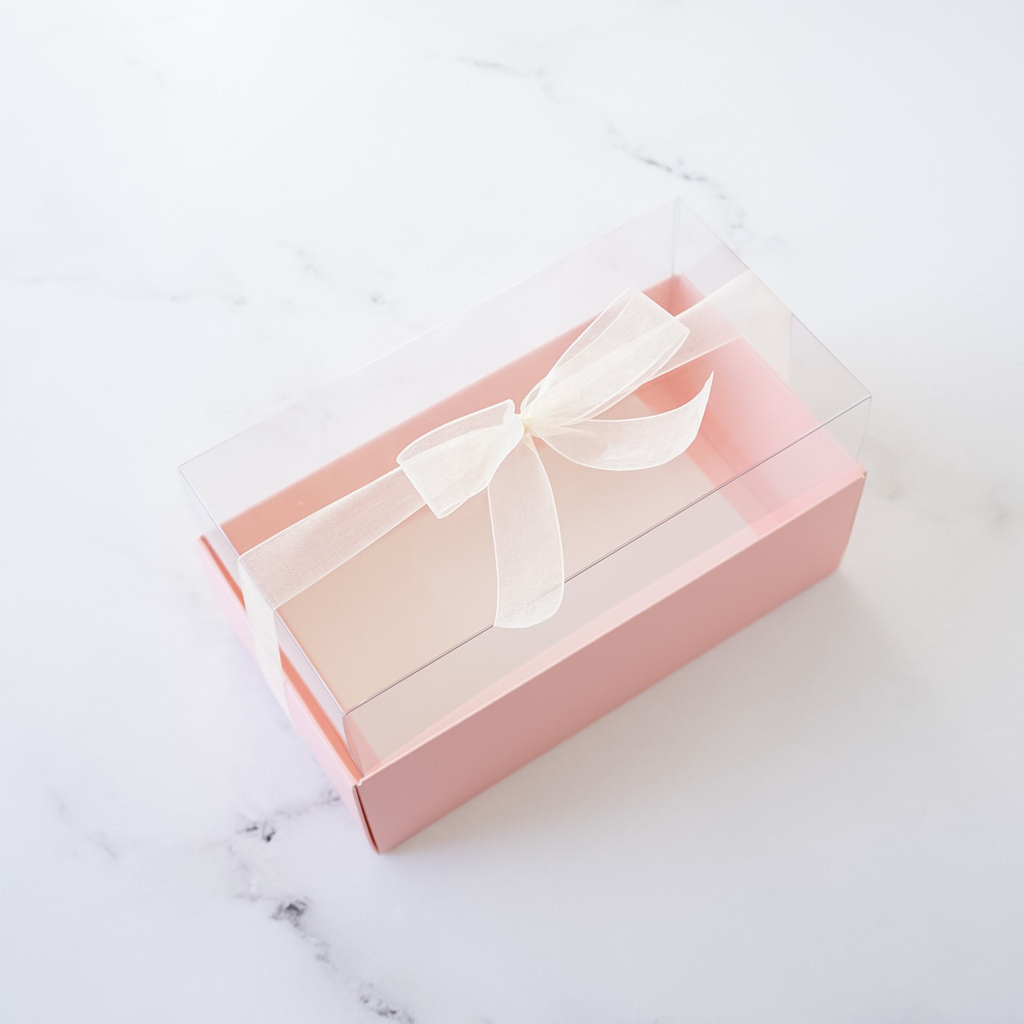 A pink gift box with a clear plastic lid. A white ribbon is tied in a bow on top of the box.
