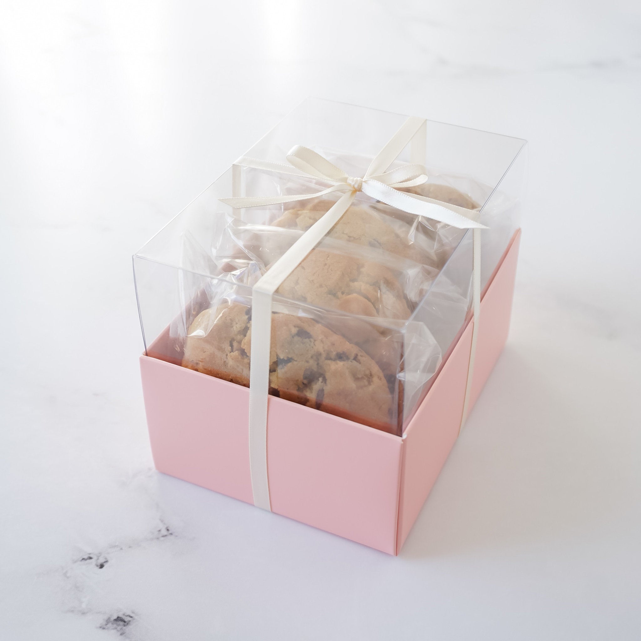 A pink box with a clear lid tied with a white ribbon. Inside the box are three cookies wrapped in clear plastic.
