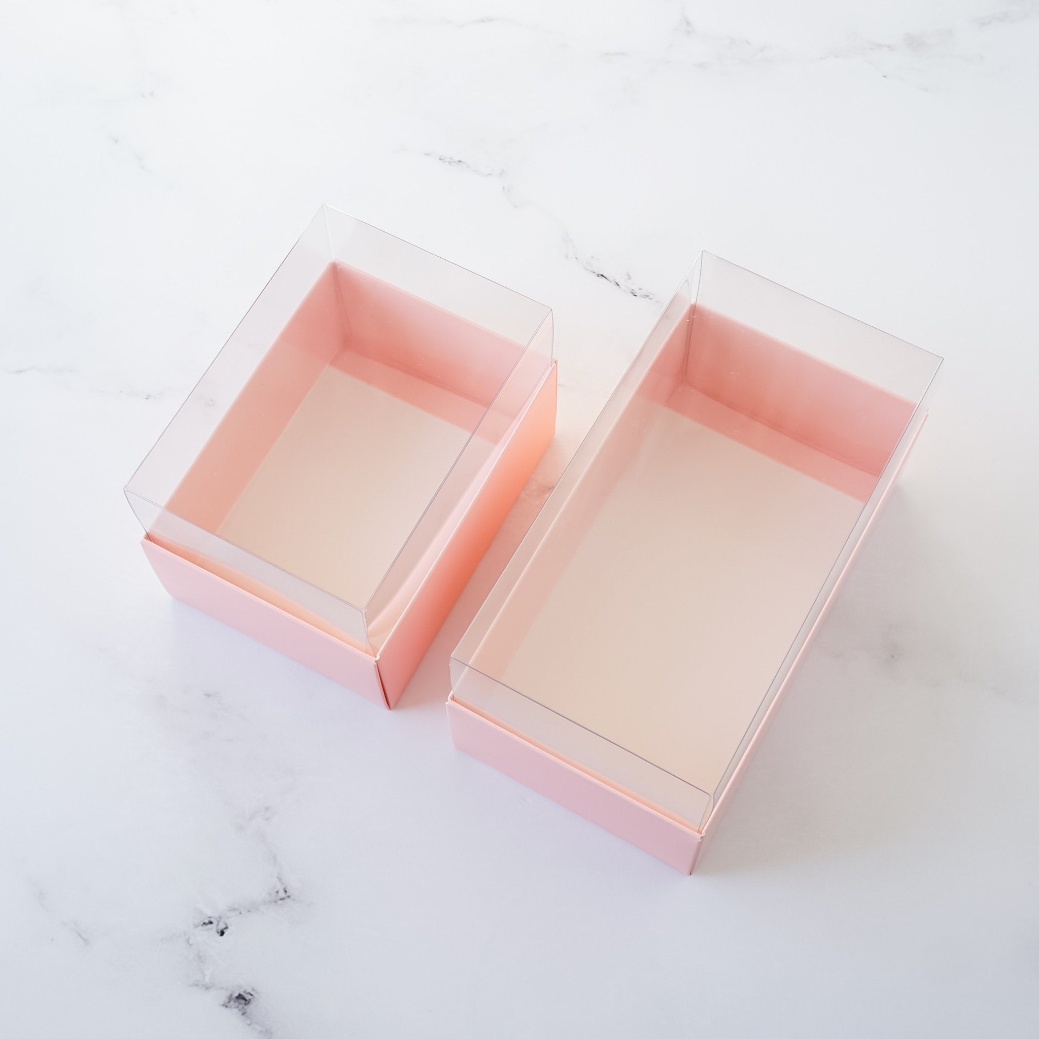 Two pink cardboard boxes with clear plastic lids. The boxes are empty and are sitting on a white marble surface.
