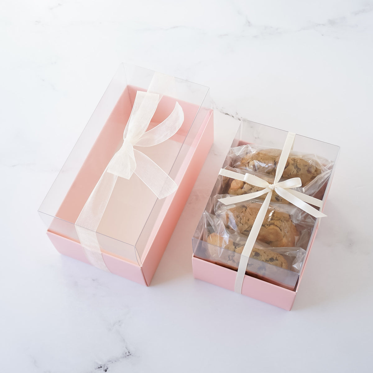 Two pink boxes with clear lids. The larger box has a white ribbon tied in a bow. The smaller box contains several cookies wrapped in clear plastic and tied with a white ribbon.