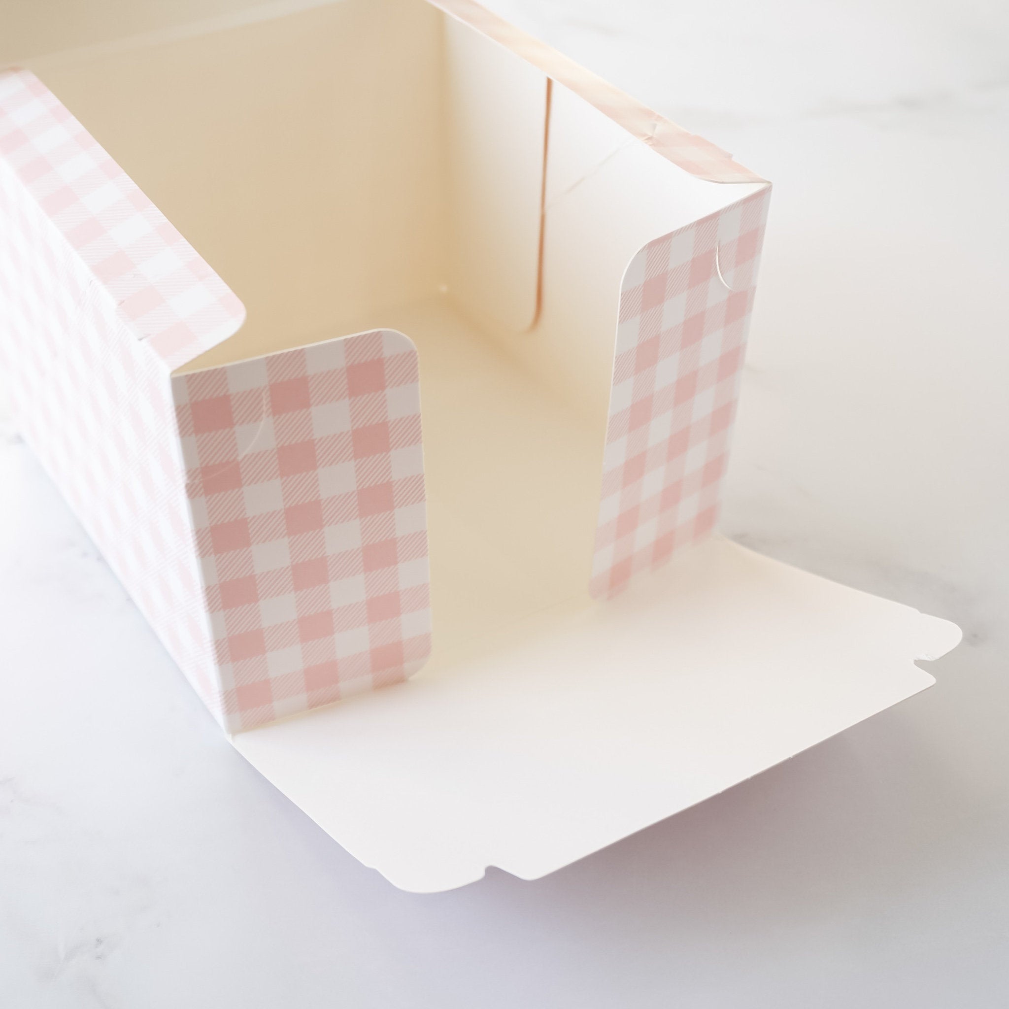 A white cardboard box with pink and white gingham pattern on the sides. The box is open and the inside is visible. The box is designed for packaging baked goods.