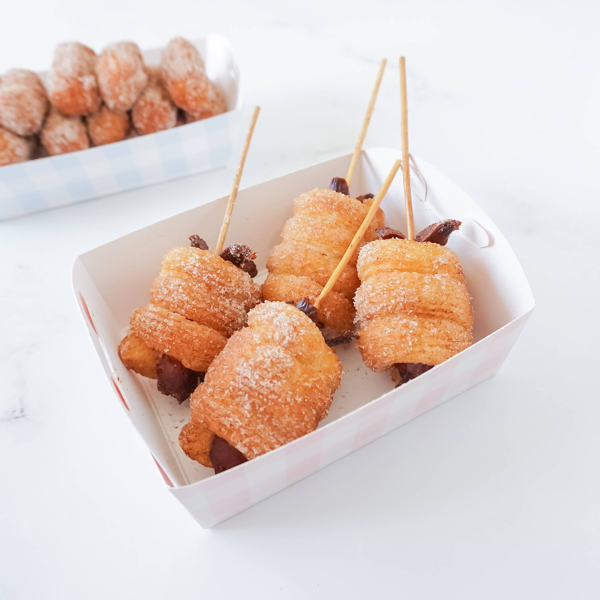 Four cinnamon sugar churros on sticks with chocolate filling in a white and pink checkered paper container