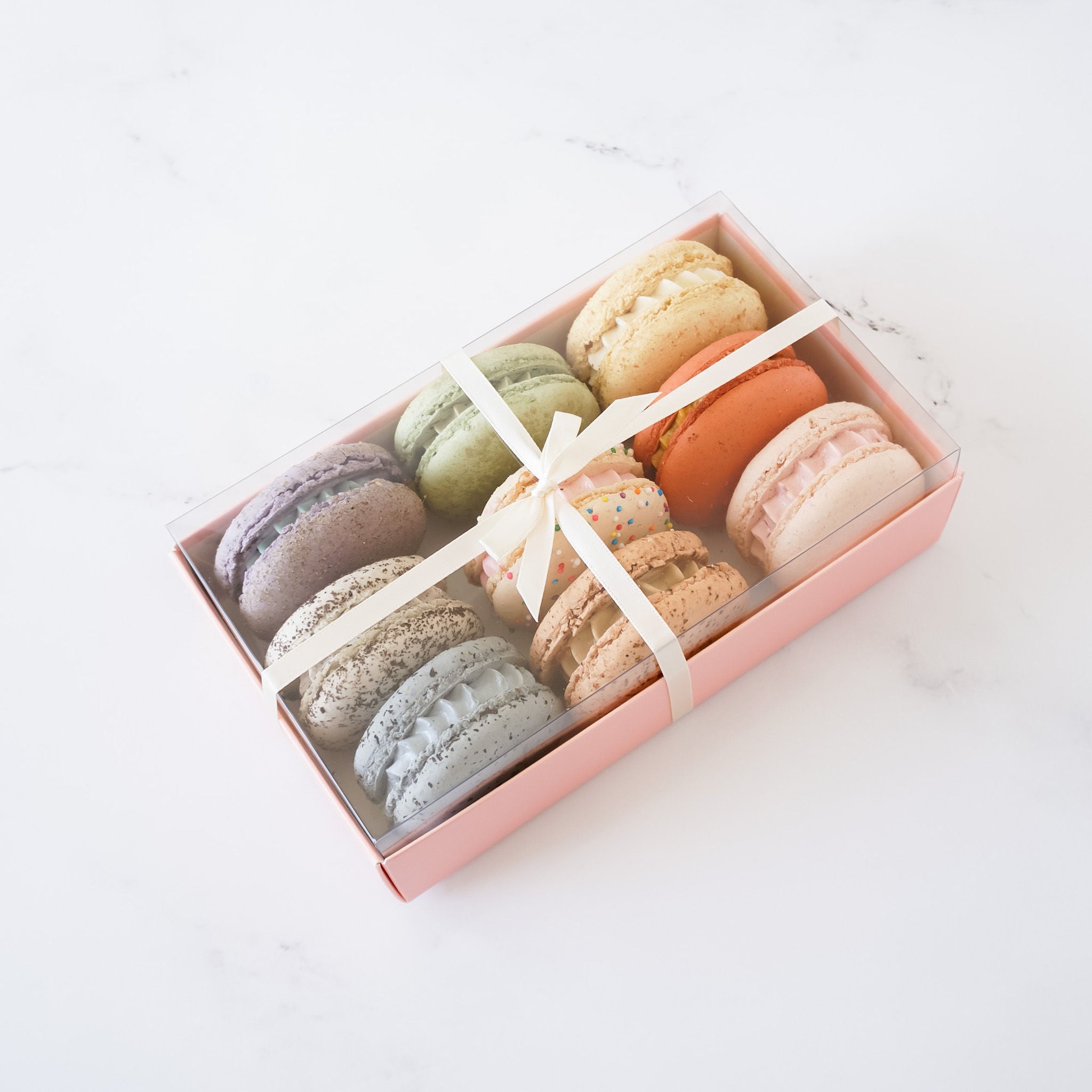 A pink box with a clear lid filled with nine colorful macarons. The macarons are arranged in three rows of three. The macarons are a variety of colors, including pink, green, blue, and purple. The box is tied with a white ribbon.