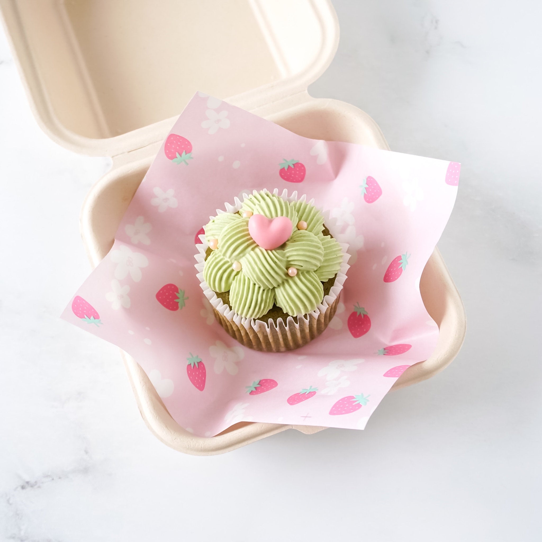 A cupcake wrapped in pink deli paper with a strawberry print, placed inside a biodegradable container.