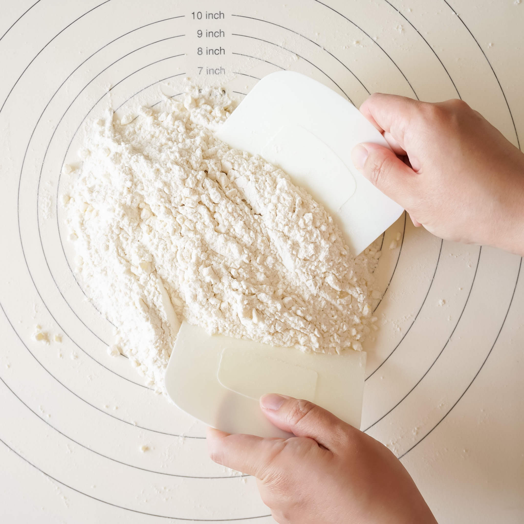 Two white plastic dough scrapers on a white surface with concentric circles printed on it. The surface has markings for 7, 8, 9, and 10 inches.