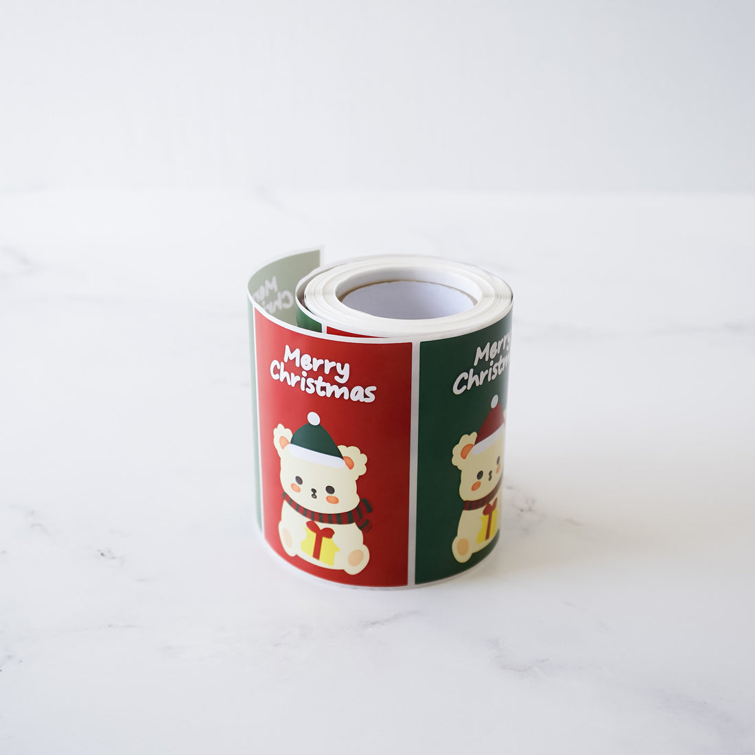 A roll of Christmas gift stickers features adorable cartoon bears wearing Santa hats. The stickers are predominantly red and green, with the phrase &