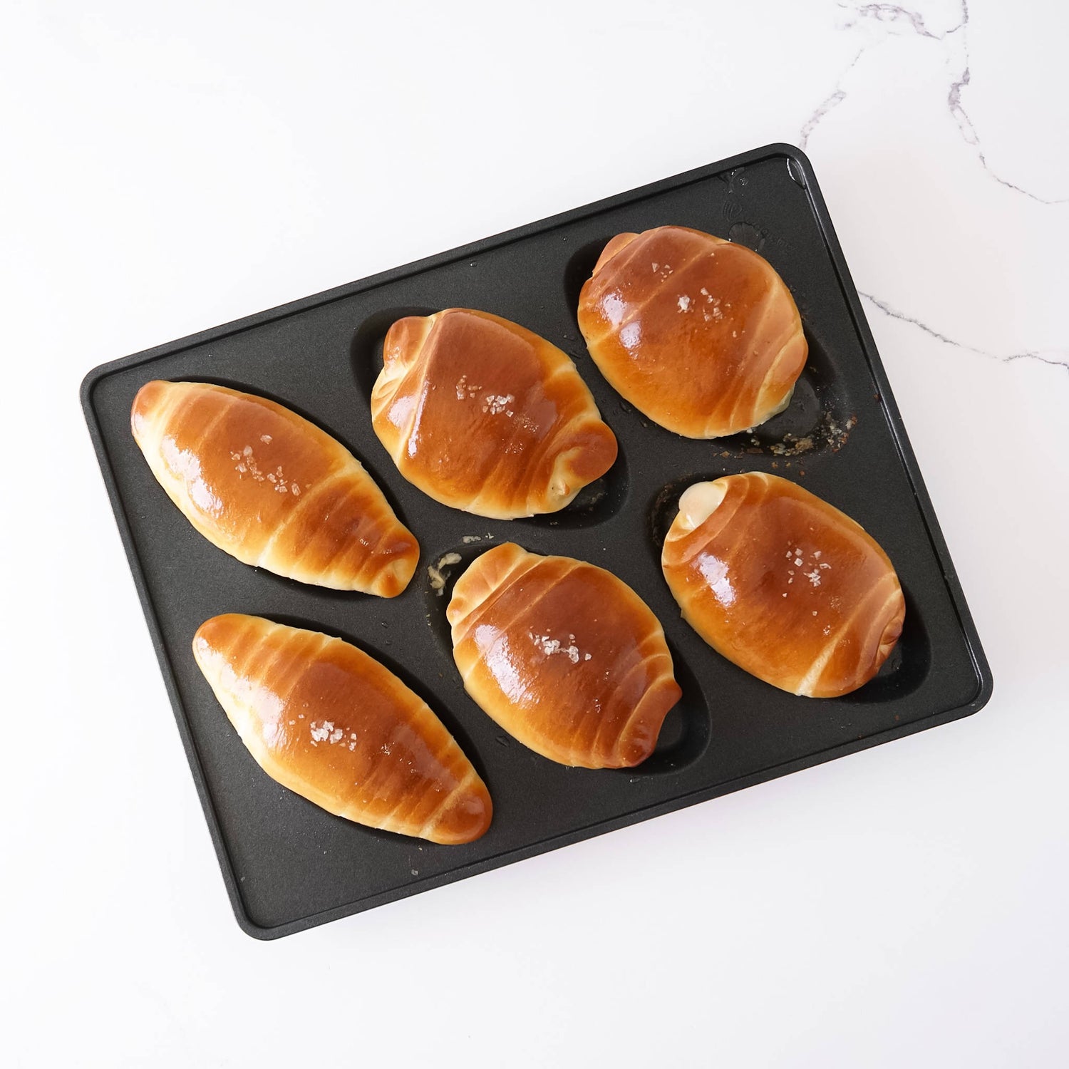Six golden-brown baked salt bread rolls arranged in a black rectangular baking tray on a white marble surface.