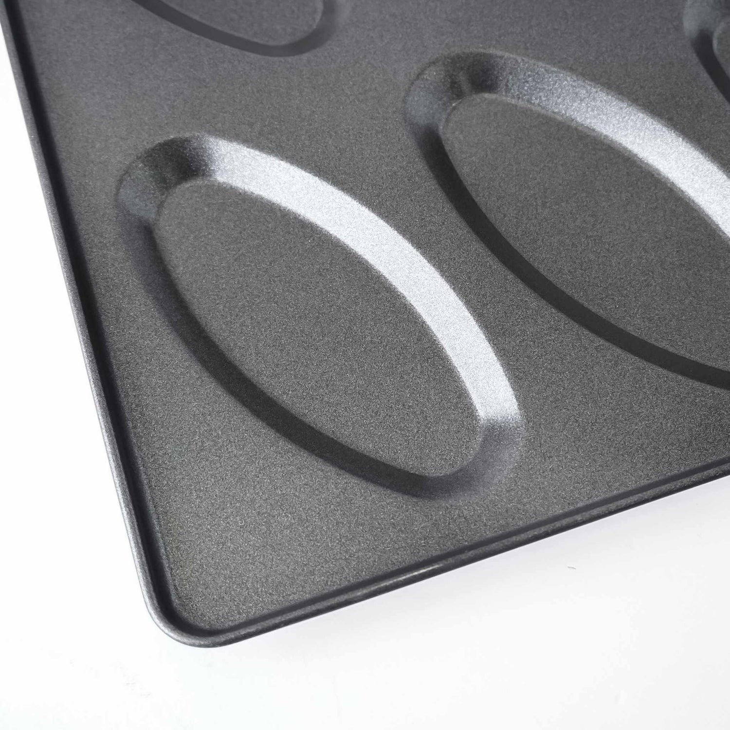 Close-up of a dark gray non-stick baking pan with oval-shaped molds, showing the textured surface and raised edges on a white background.