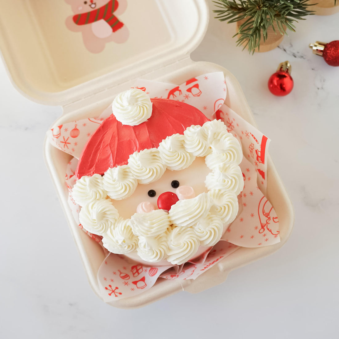 A Santa Claus-shaped Christmas cake in a takeout container. The cake is decorated with red and white frosting, creating a Santa hat and beard.  The cake&