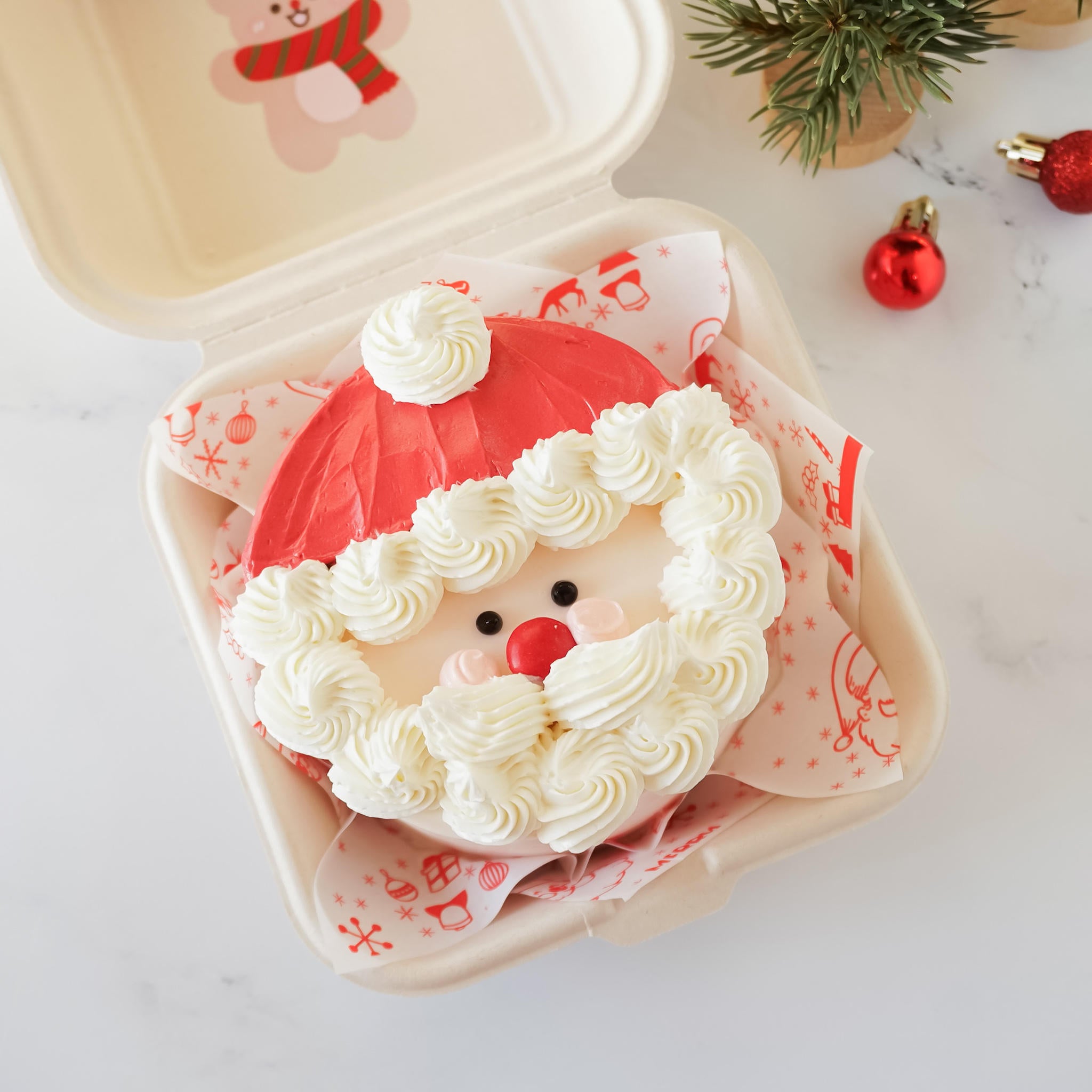 A Santa Claus-shaped Christmas cake in a takeout container. The cake is decorated with red and white frosting, creating a Santa hat and beard.  The cake&