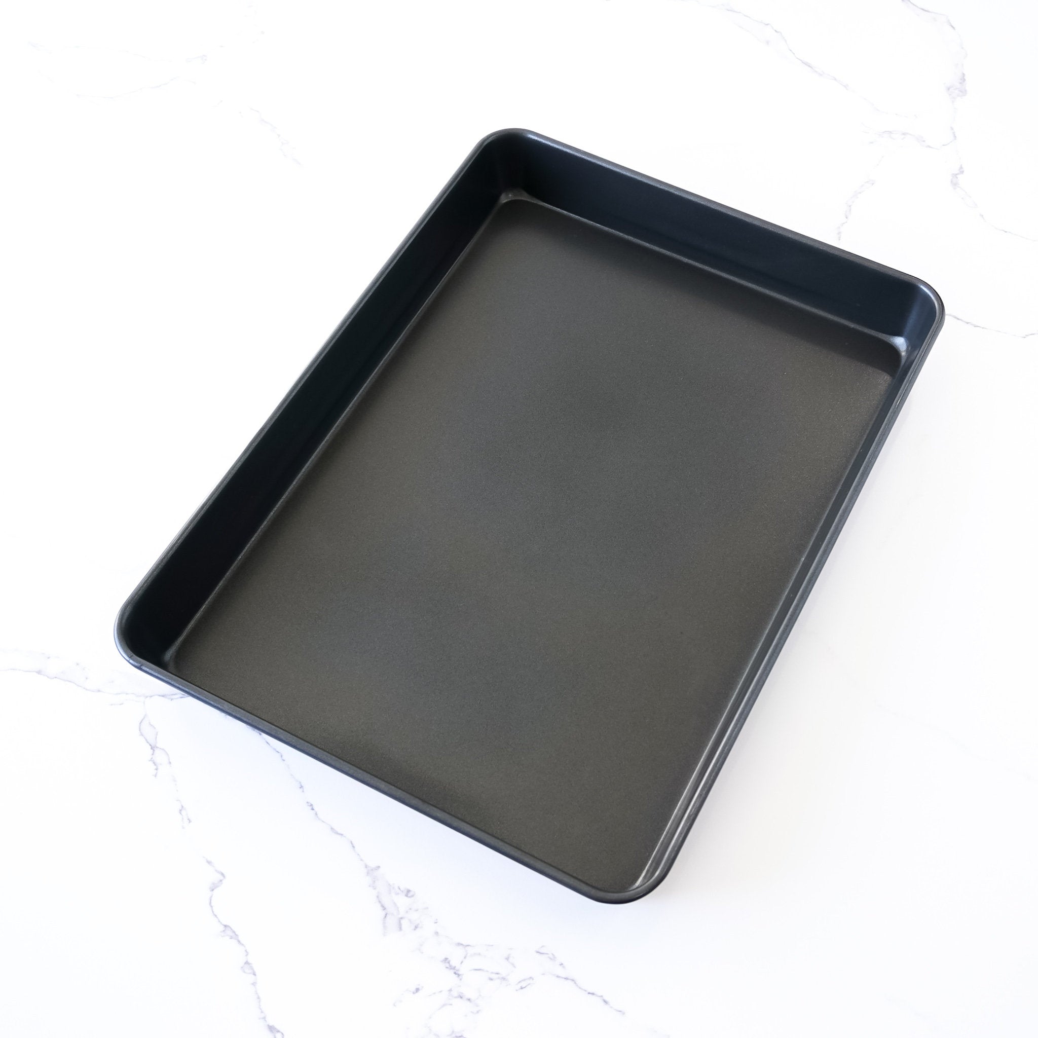A rectangular, black sheet cake pan made of nonstick coating material, placed on a white marble surface.