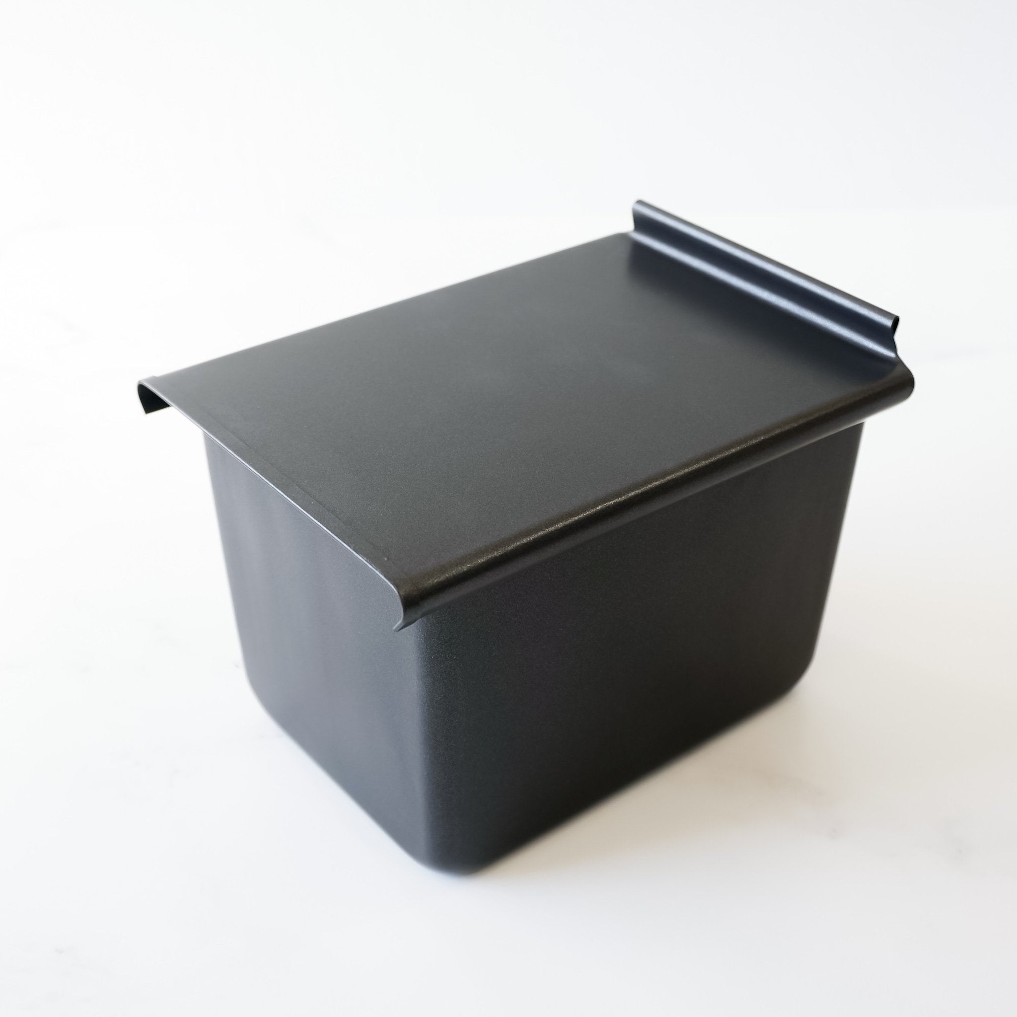 A black rectangular metal container with a lid. The container has a slightly rounded edge on the top and bottom. The lid is slightly angled and has a rounded edge on the top.