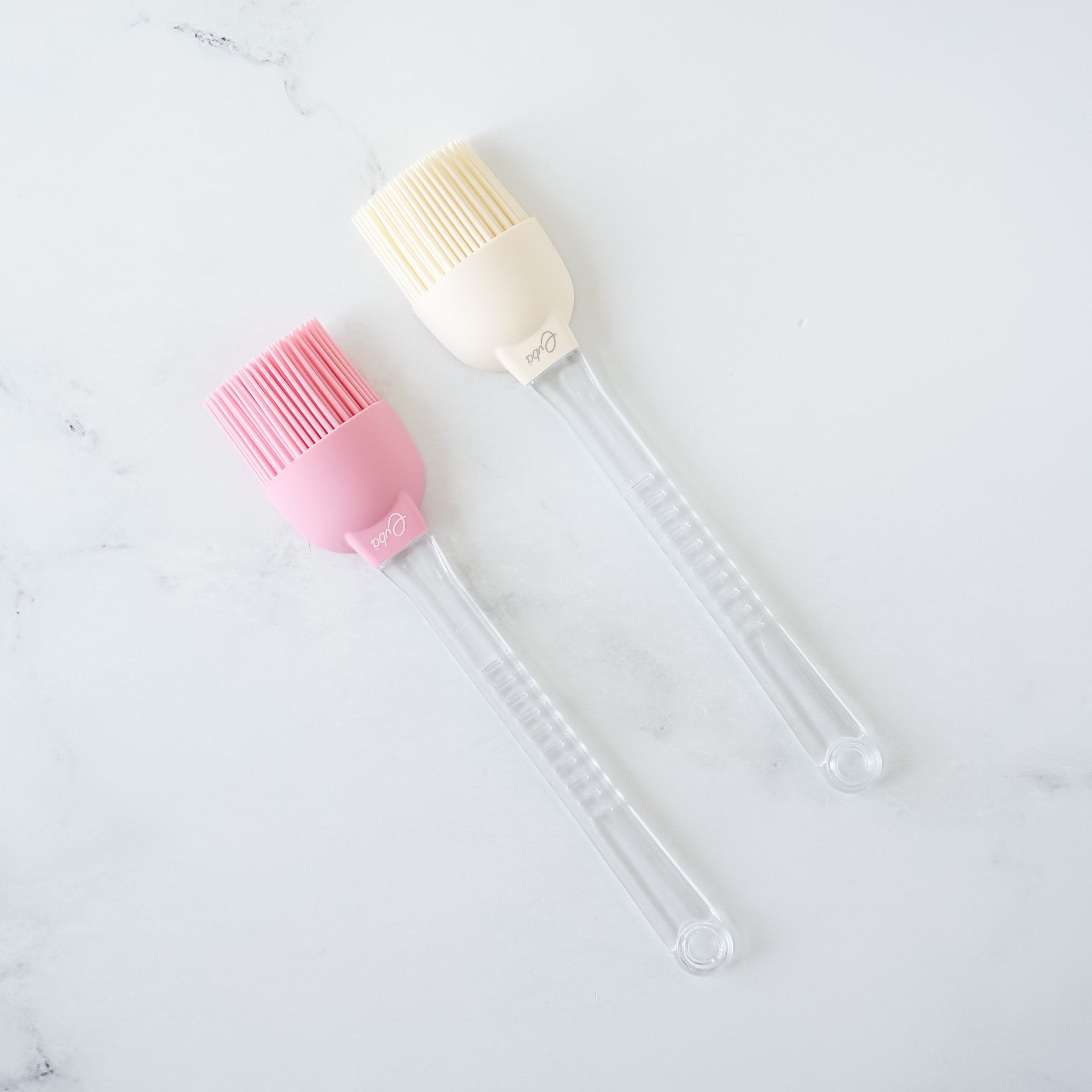 Two silicone basting brushes with detachable plastic handles, one in ivory and one in pink, on a marble surface.