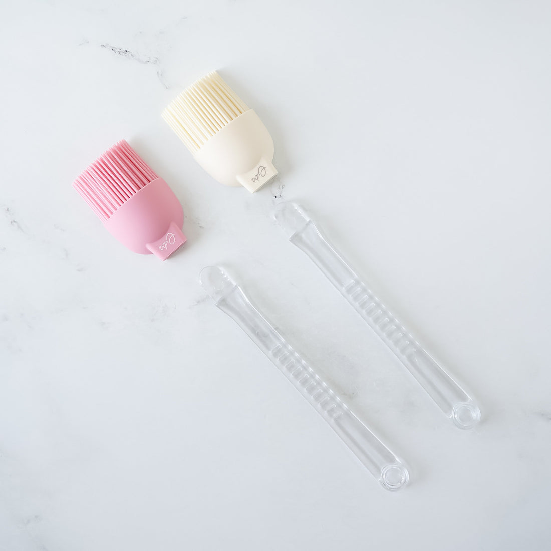 Two silicone basting brushes with detachable plastic handles, one in ivory and one in pink, on a marble surface.