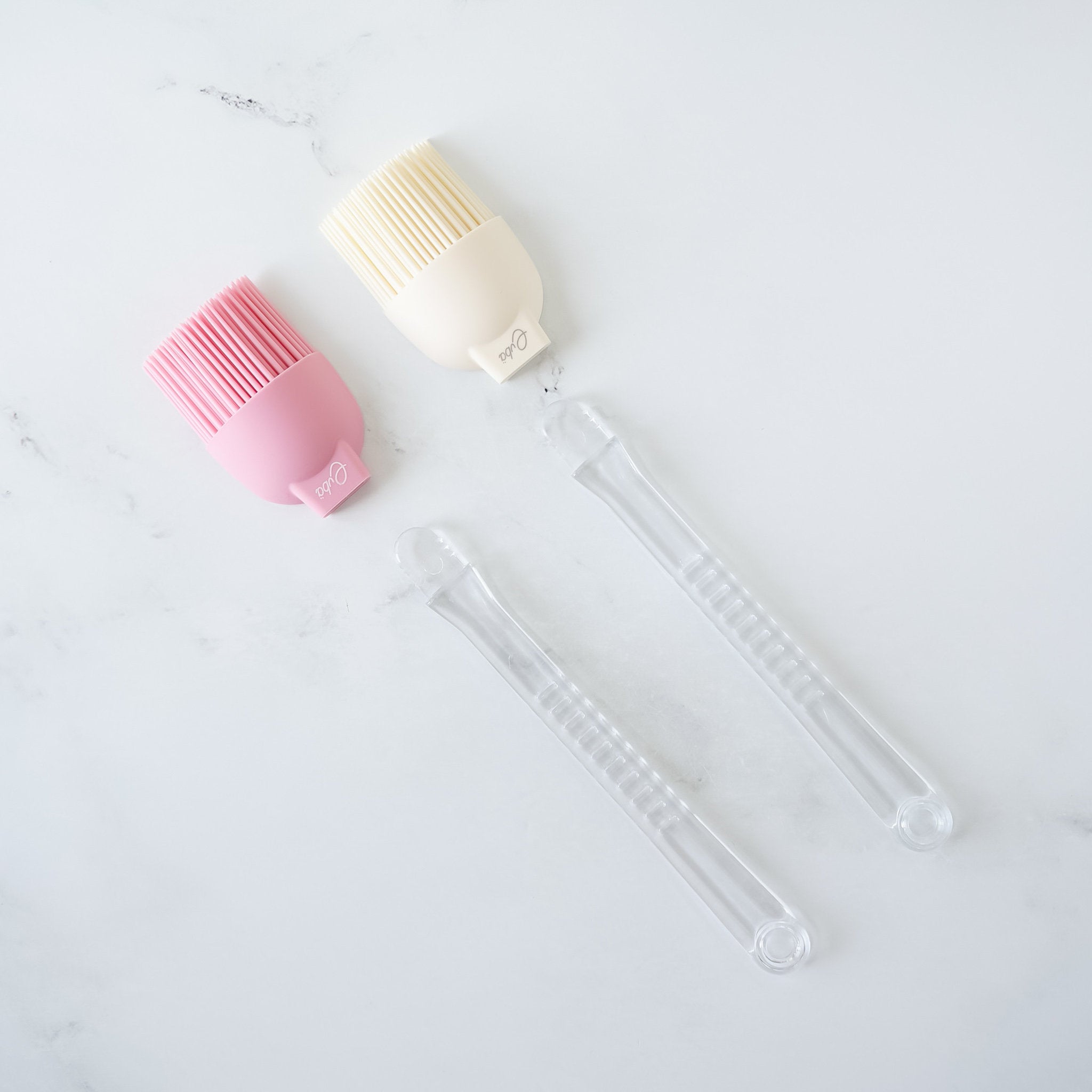 Two silicone basting brushes with detachable plastic handles, one in ivory and one in pink, on a marble surface.