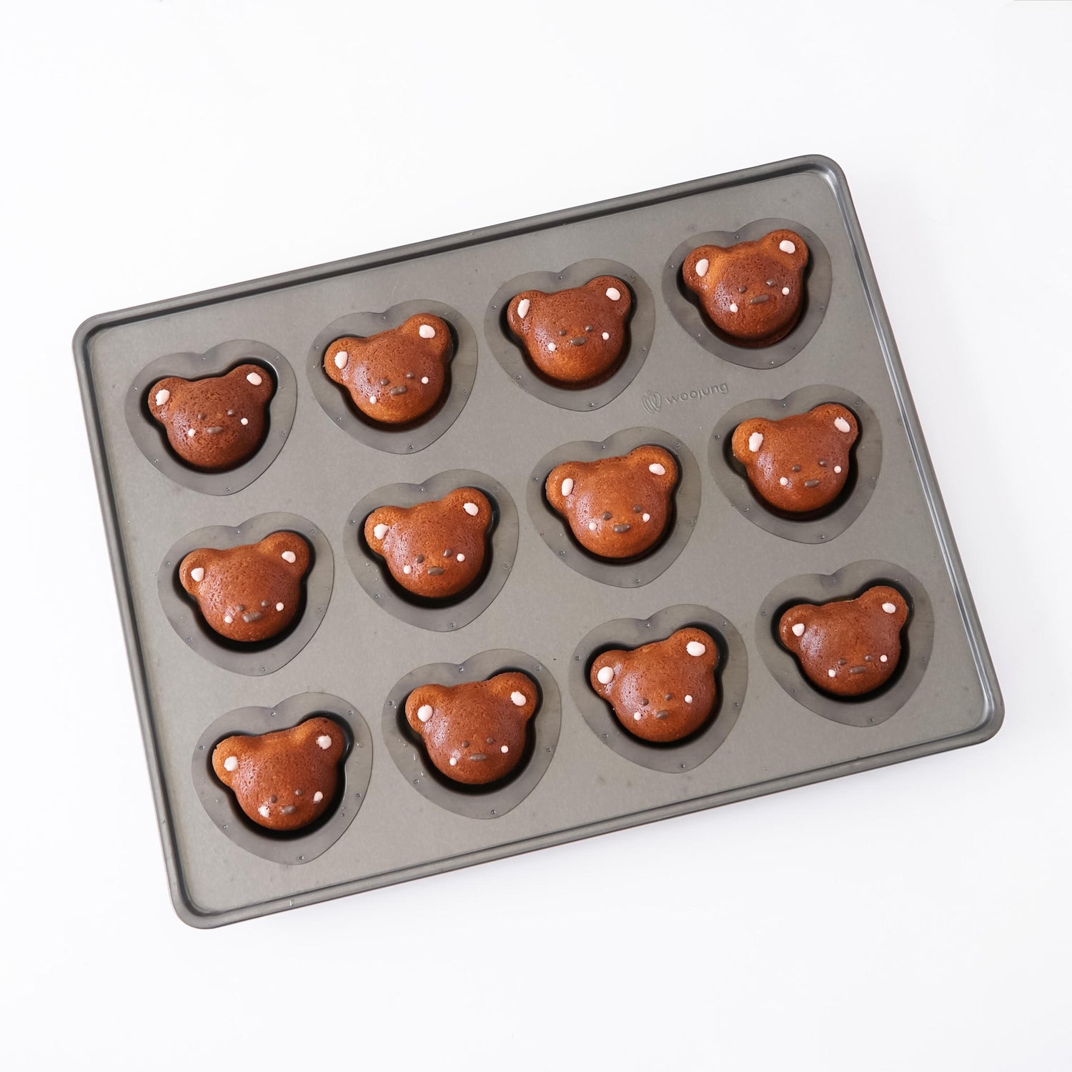 A dark gray silicone coated mold tray with twelve bear-shaped cavities, shown partially filled with baked brown bear-shaped madeleines on a white background.
