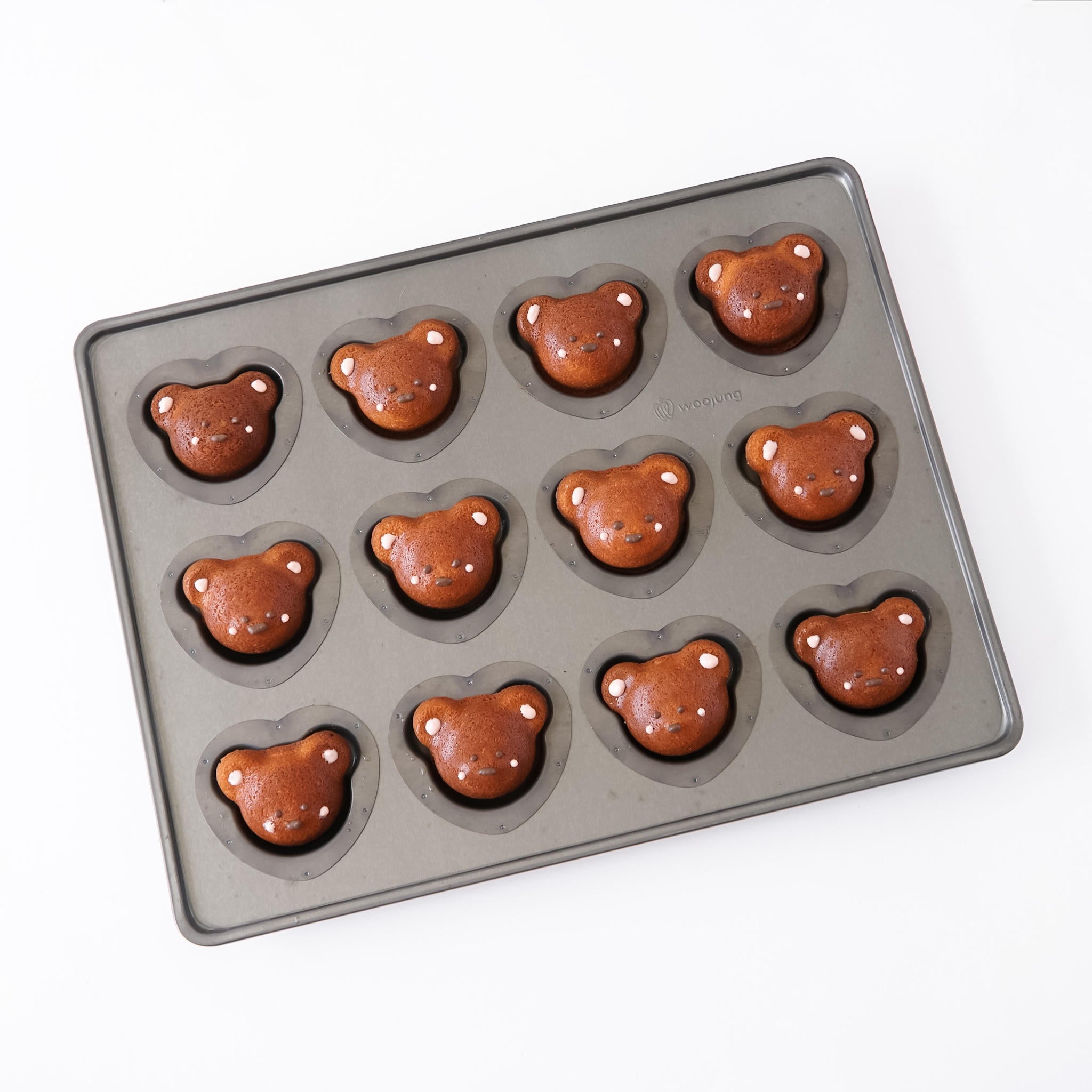 A dark gray silicone coated mold tray with twelve bear-shaped cavities, shown partially filled with baked brown bear-shaped madeleines on a white background.