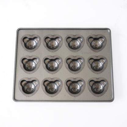 A rectangular dark gray metal baking pan with twelve bear-shaped cavities, each containing a teddy bear face mold, shown on a plain white background.
