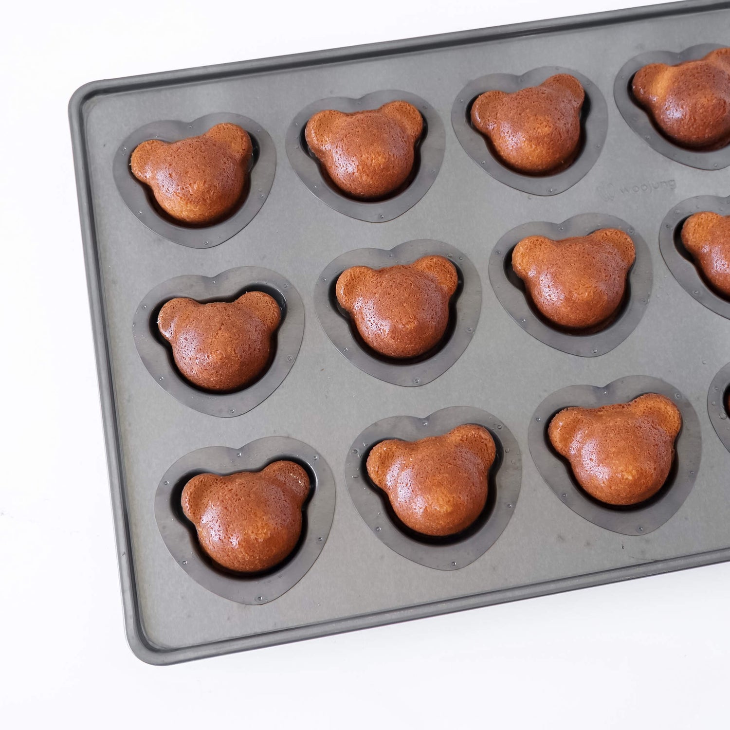 A dark gray silicone coated mold tray with bear-shaped cavities, each containing a baked brown madeleine cake shaped like a bear head, shown on a white background.