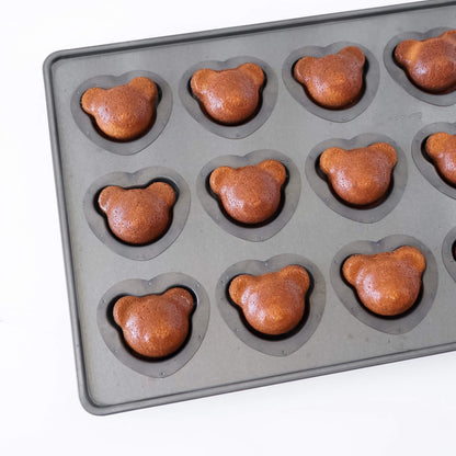 A dark gray silicone coated mold tray with bear-shaped cavities, each containing a baked brown madeleine cake shaped like a bear head, shown on a white background.