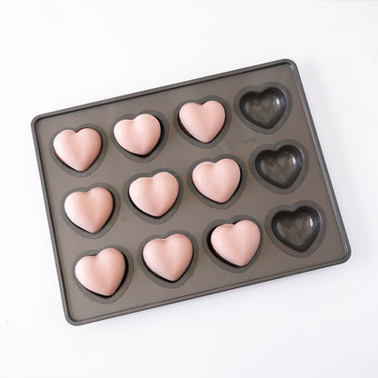 A rectangular dark gray silicone mold tray with 12 heart-shaped cavities, some filled with pale pink chocolate cake and others empty, shown on a white background.