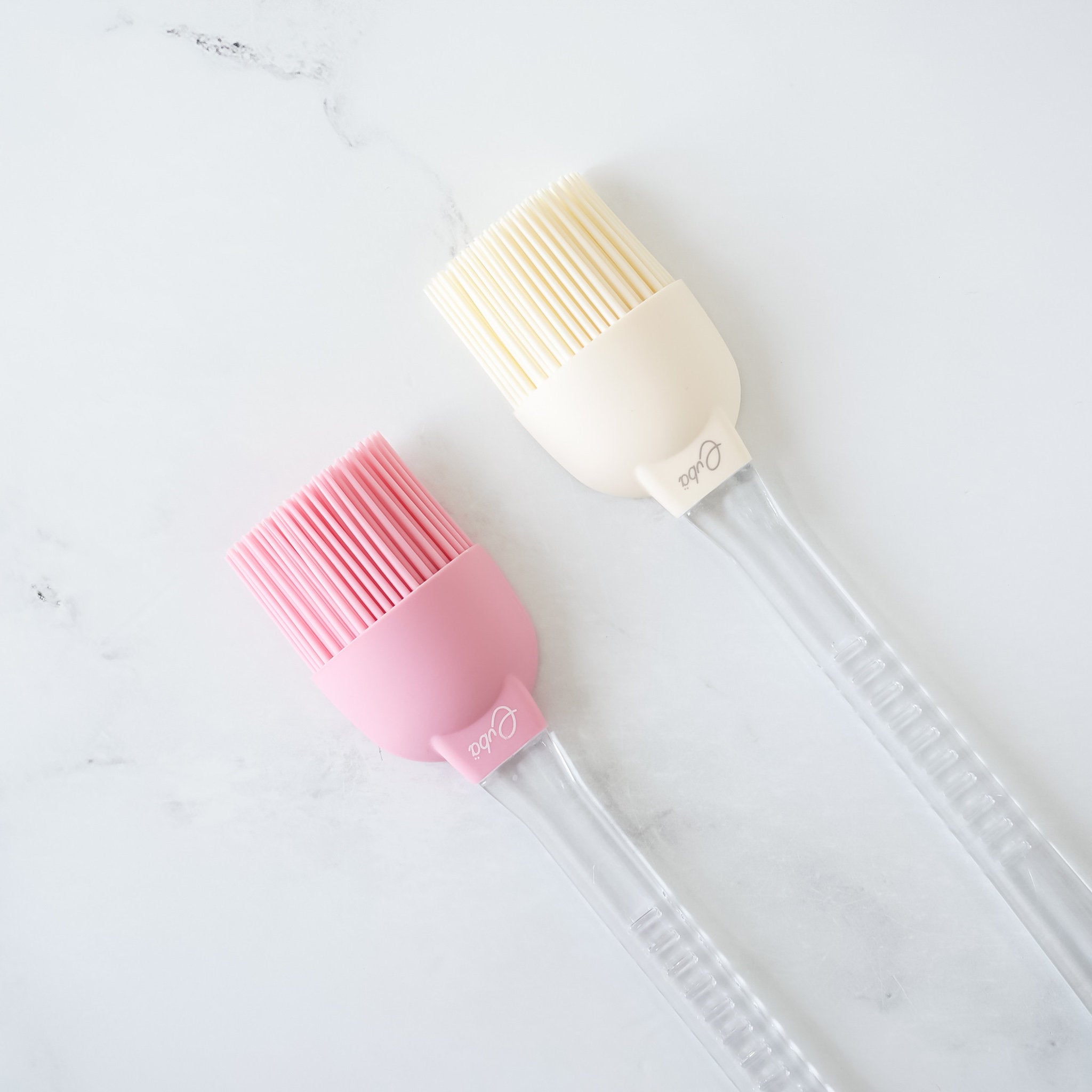 Two silicone basting brushes with detachable plastic handles, one in ivory and one in pink, on a marble surface.