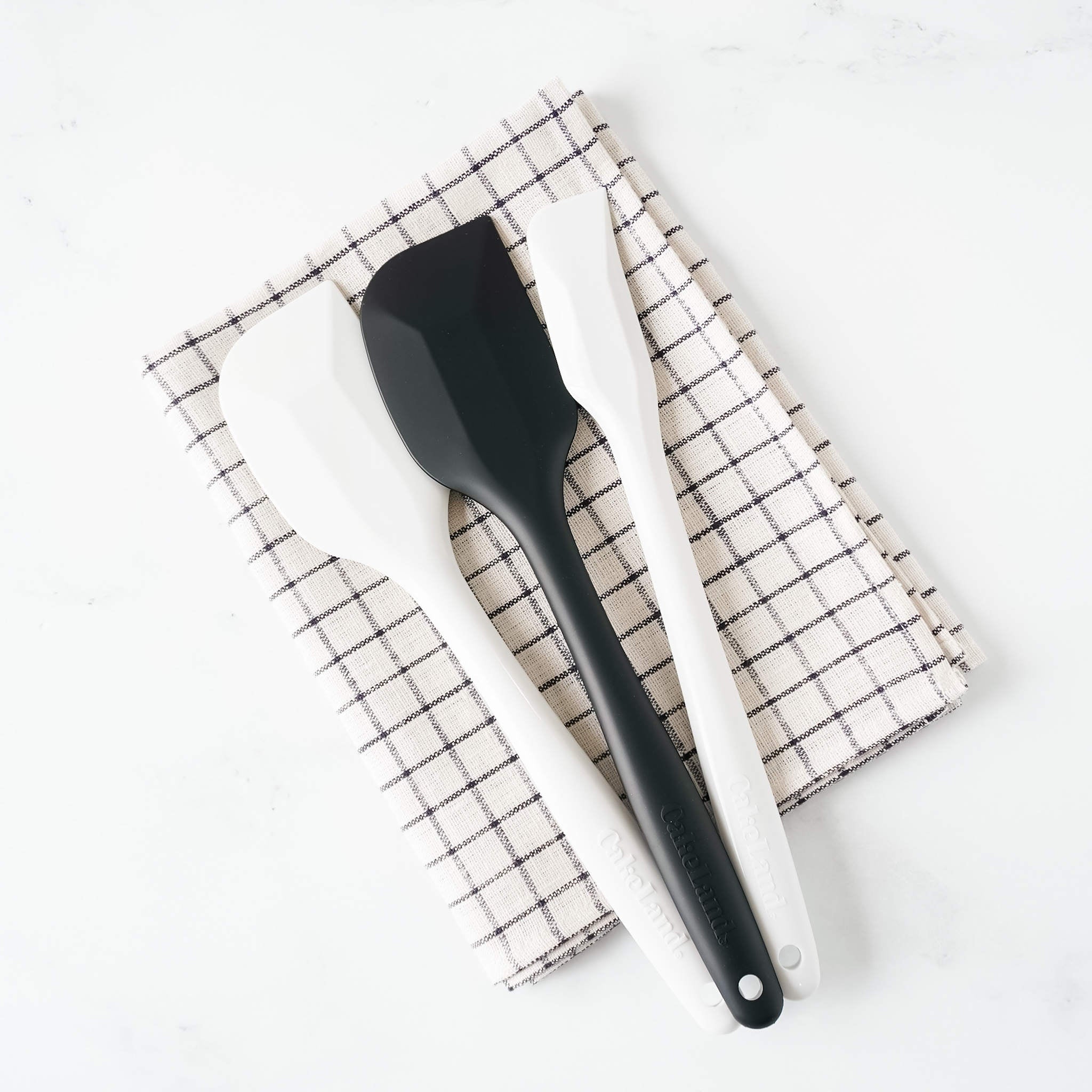 A black and two white silicone spatulas by Tiger Crown placed on a checkered napkin.