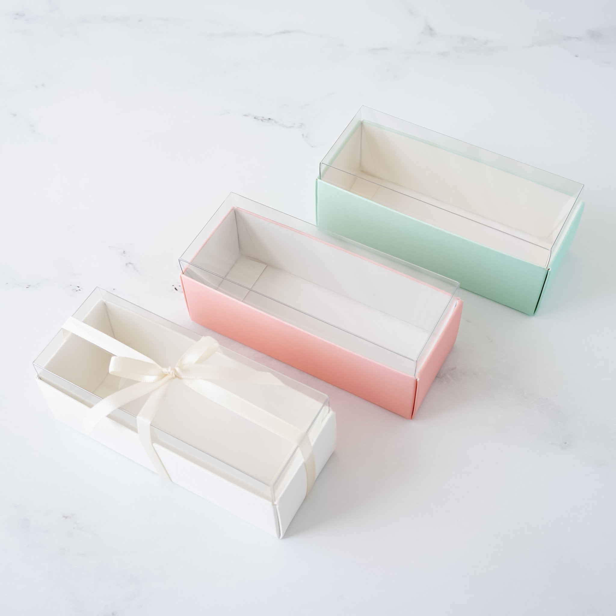 Three rectangular boxes with clear lids. The boxes are empty and are arranged in a row from left to right. The box on the left is white, the middle box is pink, and the box on the right is light green.