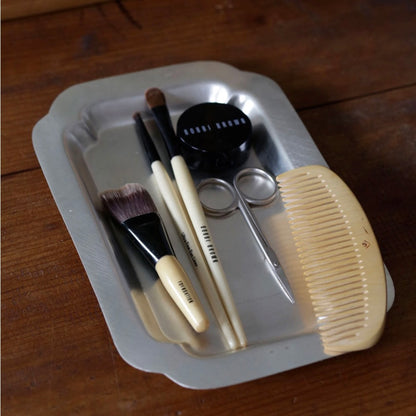 A set of hair styling tools and accessories arranged in a small silver tray on a wooden table, including a beige comb, makeup brushes, metal scissors, and a black round container.