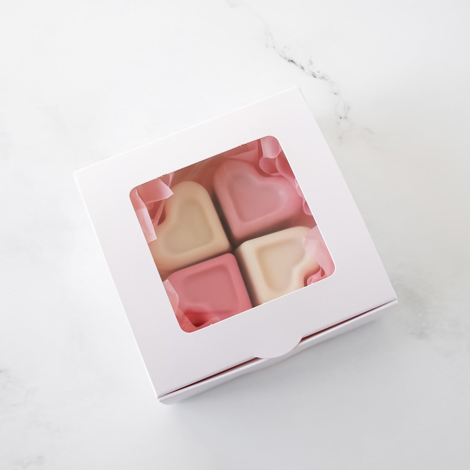Four pink and white square candies in a white bakery box on a white marble surface.