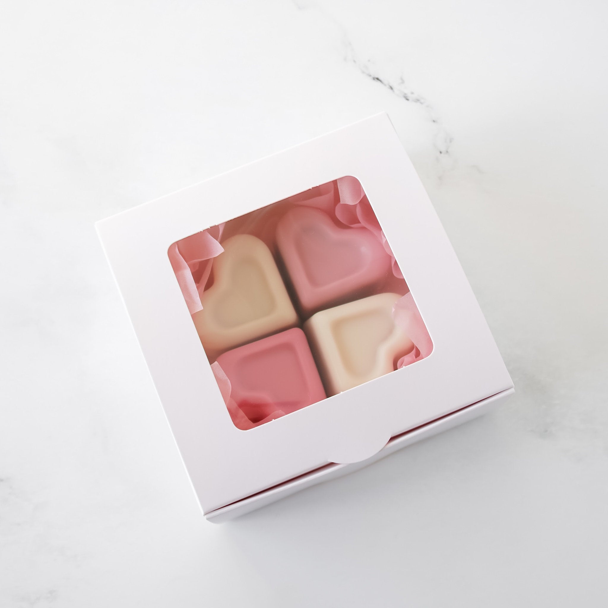 Four pink and white square candies in a white bakery box on a white marble surface.