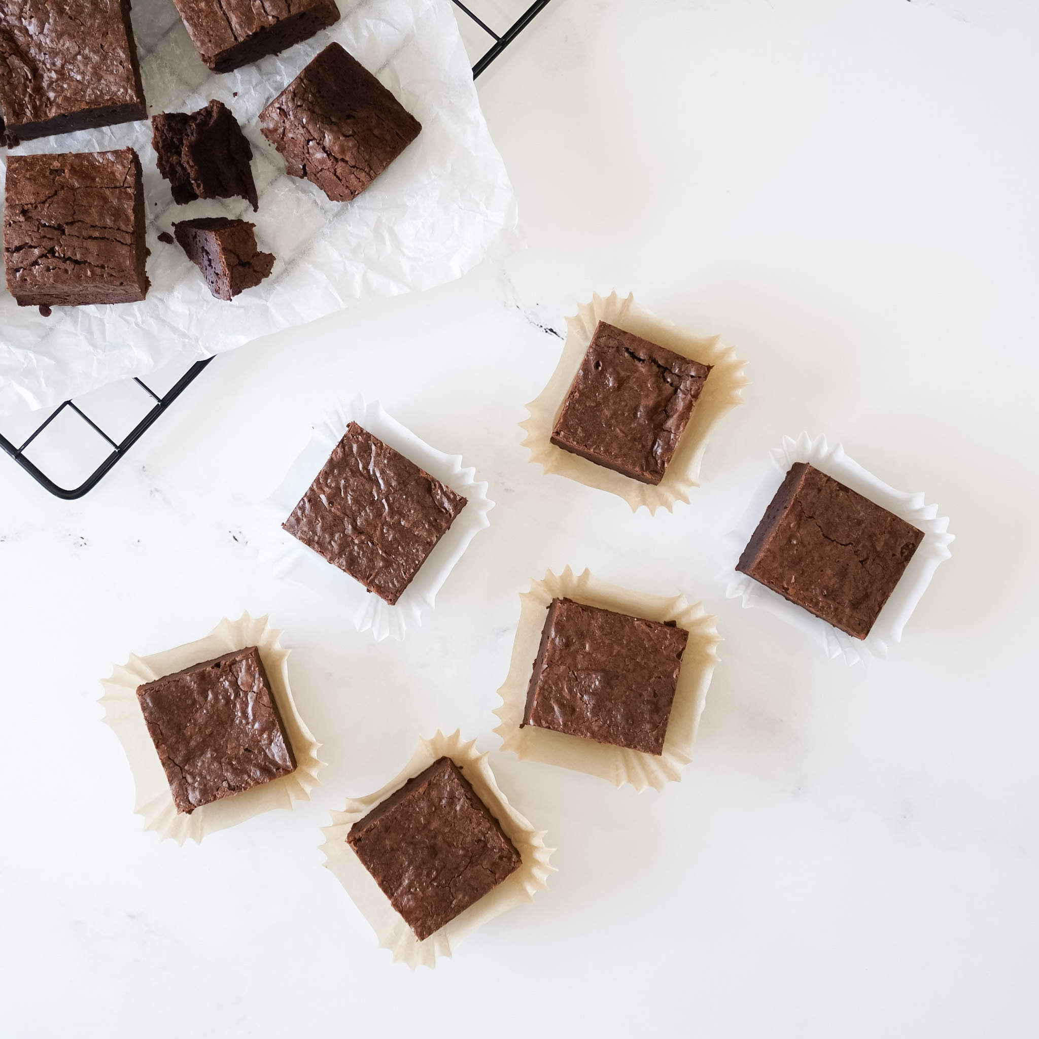 Six squares of chocolate brownies on white parchment paper. The brownies are arranged in a semi-circle. Each brownie is in a white paper wrapper.