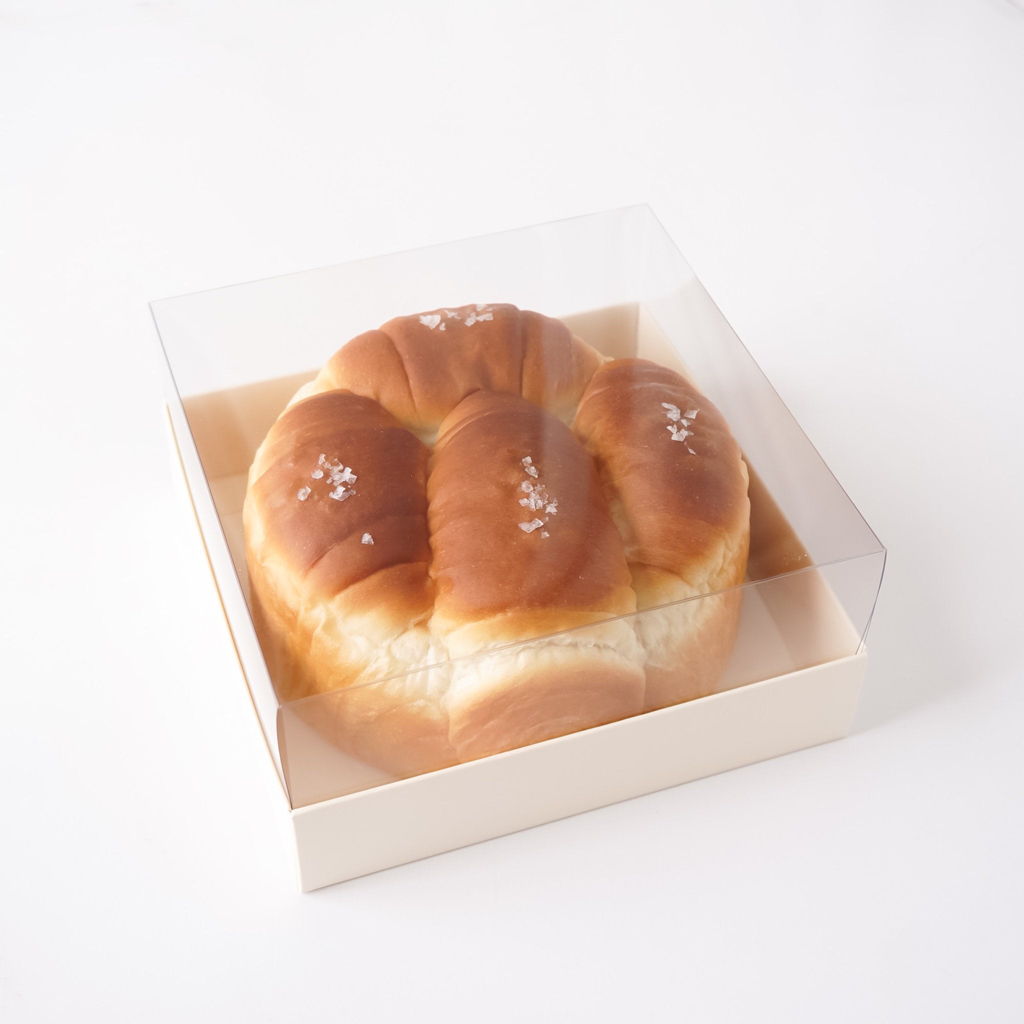 A round braided bread loaf, likely a challah or sweet bread, with coarse sea salt sprinkles, presented inside a square cream-colored bakery box with a clear plastic lid, shot on a seamless white background.