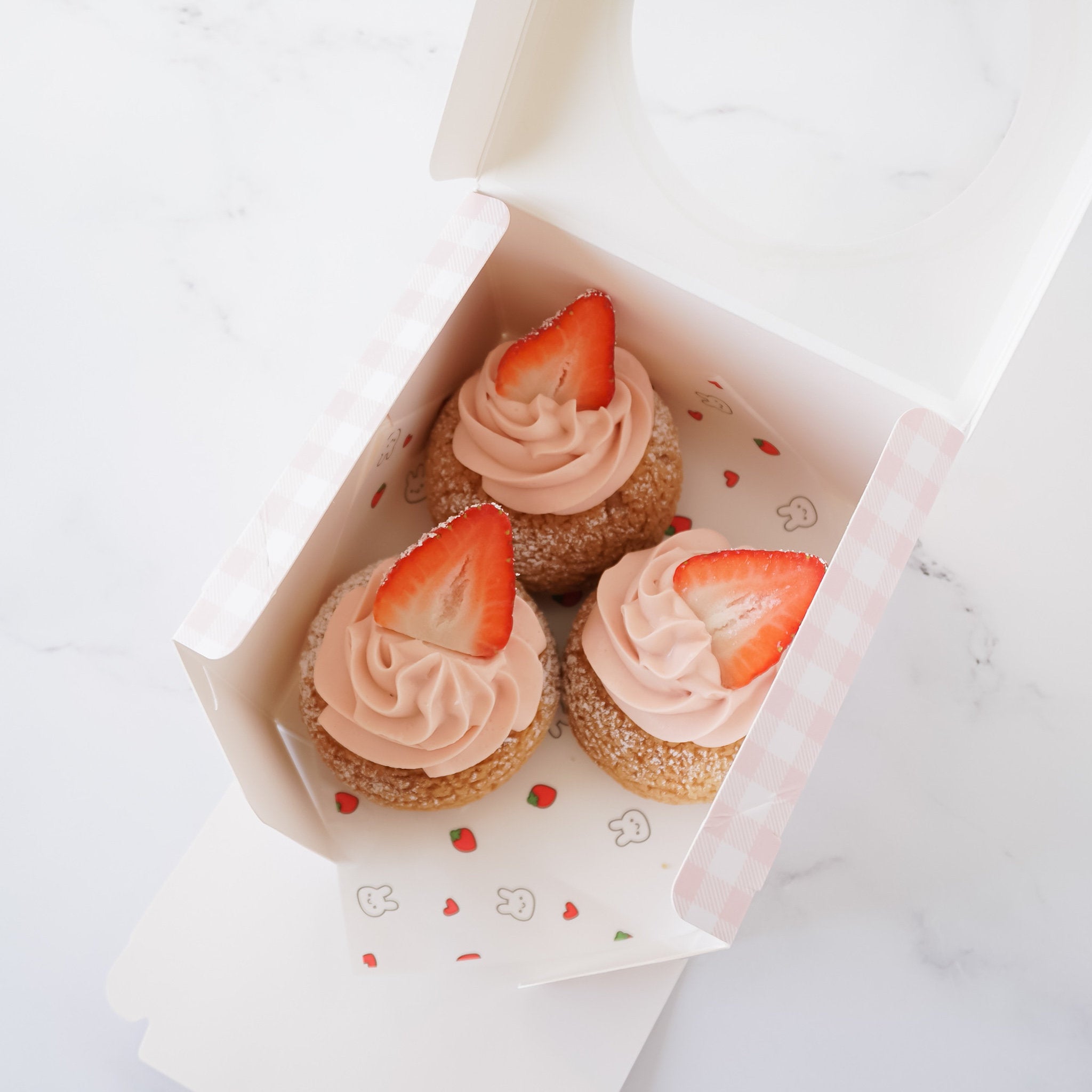 Three pink cream puffs topped with strawberries in a white and pink gingham gift box.